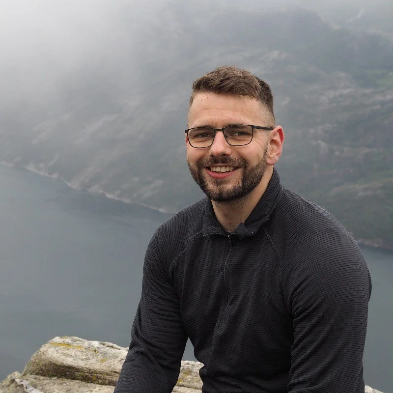 A man with glasses and a beard smiling outdoors, with a lake and mountains in the background.