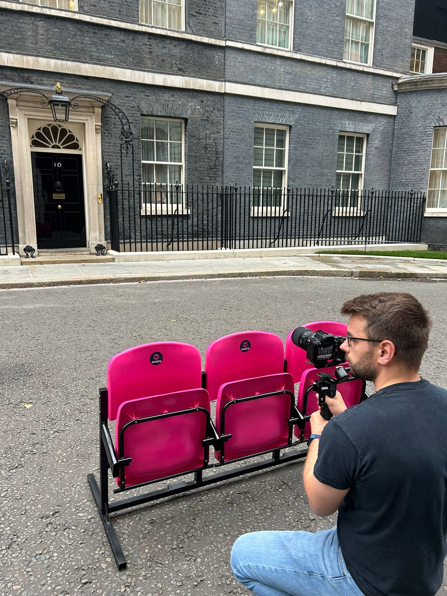 A person sitting on a pavement, holding a camera, with three pink chairs in front of them. Behind them, there is a black wrought iron fence and a large brick building with white-windowed dormers.