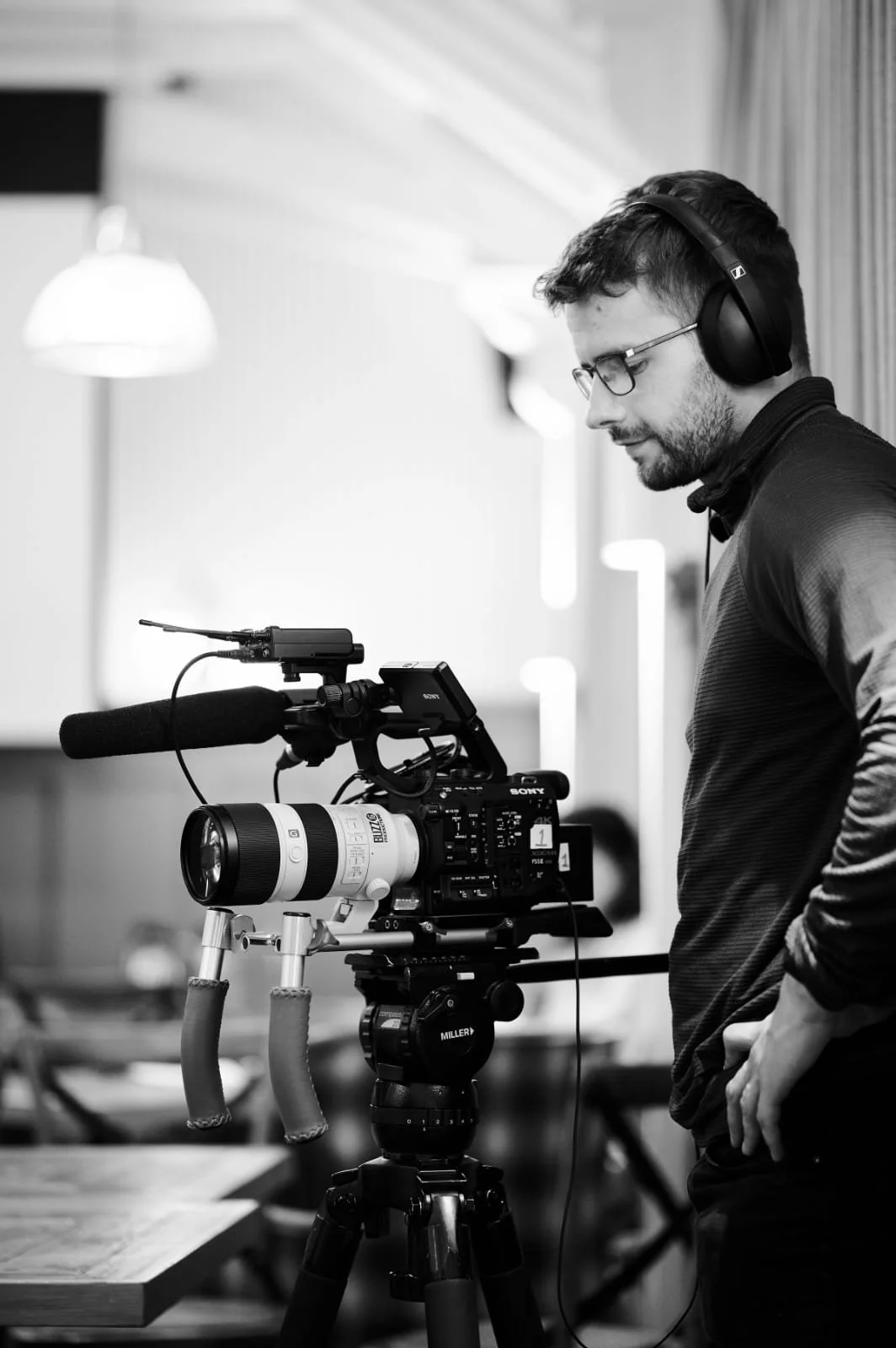 A man wearing glasses and headphones operating a professional video camera on a tripod in an indoor setting.
