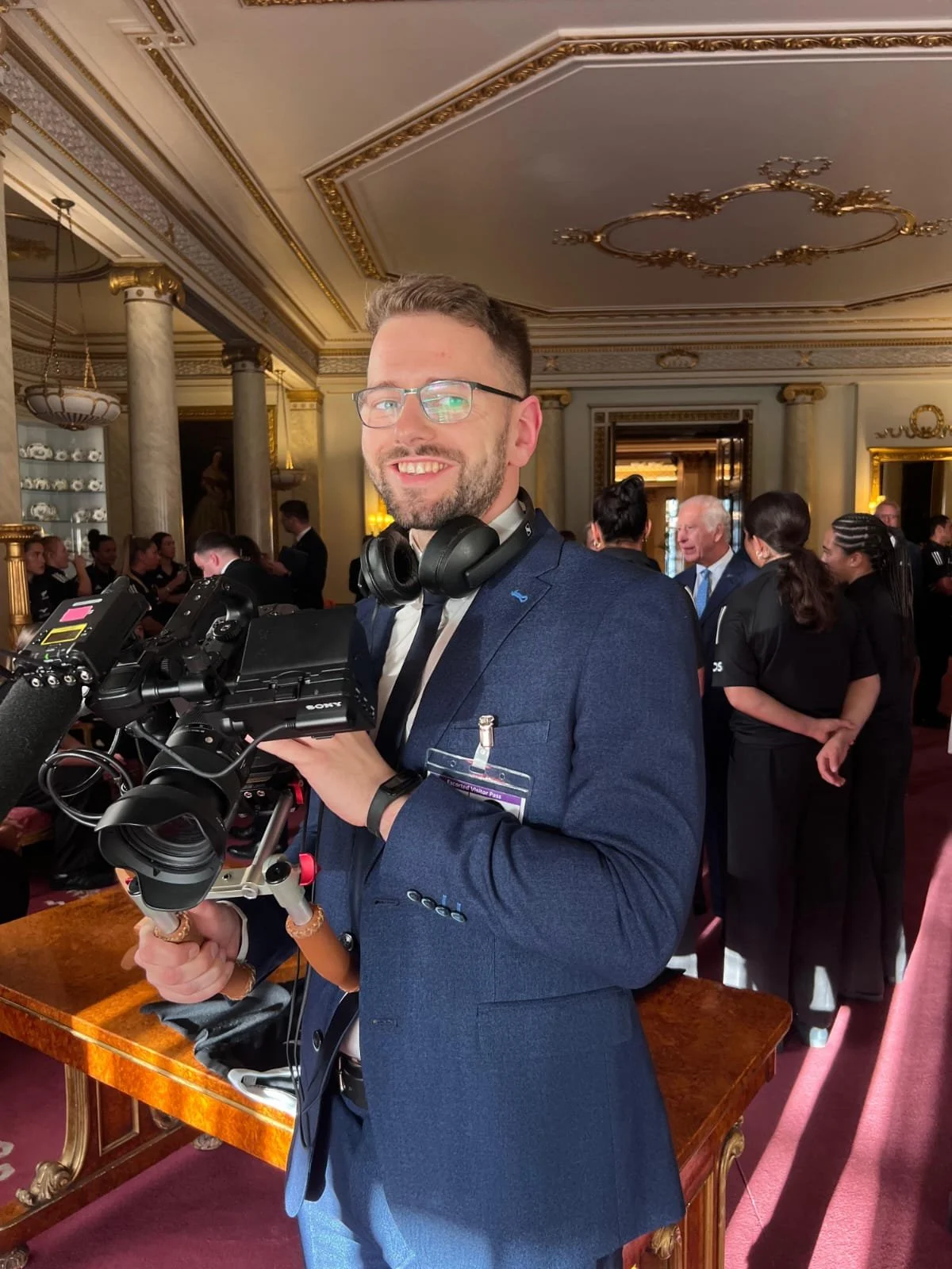 A smiling man in a blue suit holding a professional video camera. He is wearing glasses and headphones around his neck. He is at a formal event with ornate gold and marble interior decor, and people in the background are engaged in conversations.