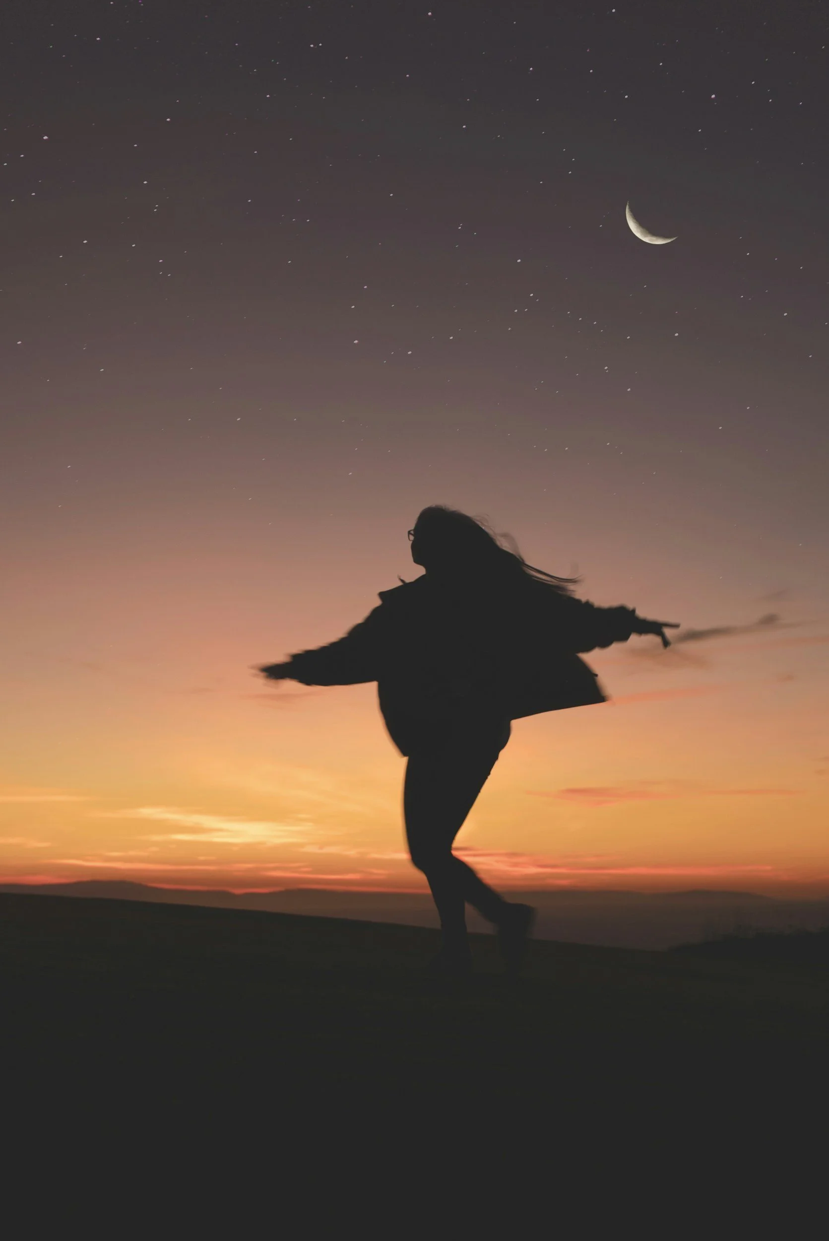 Silhouette of a person dancing or jumping outdoors during sunset with a sky full of stars and a crescent moon overhead.