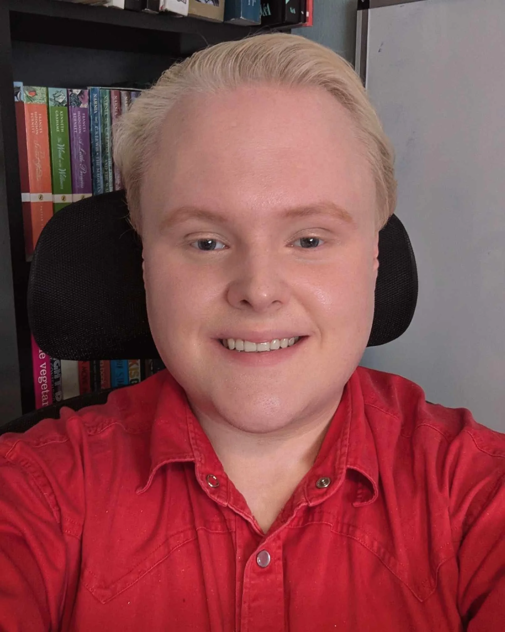 A person with blond hair smiling, wearing a red shirt, sitting in front of a bookshelf filled with colorful books.