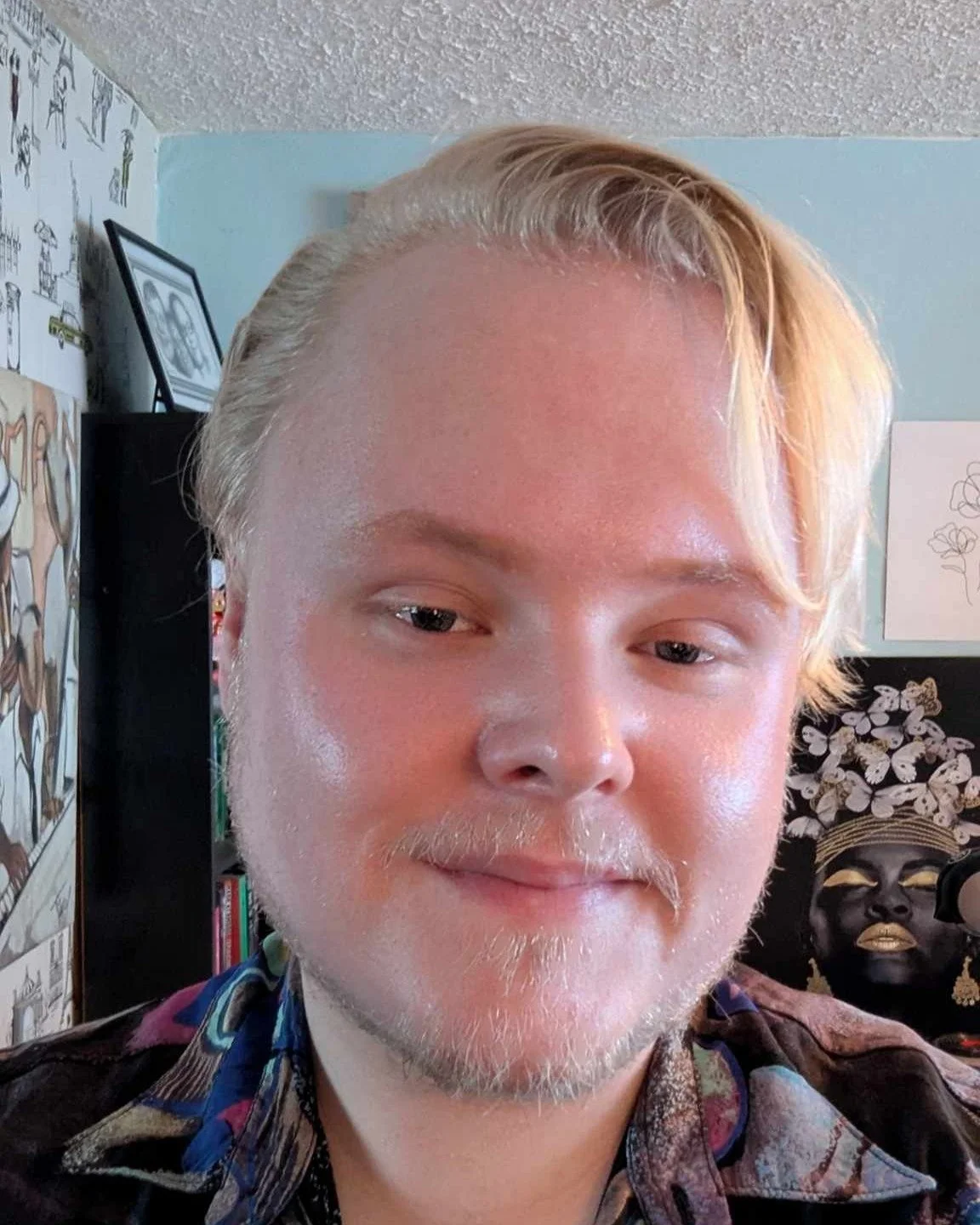 A close-up selfie of a person with light skin, blond hair, and a trimmed beard, smiling softly with slightly squinted eyes. The background features framed pictures, a bookshelf, and a dark sculpture of a face with flowers on its head.