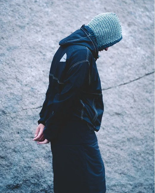 Person in a black jacket and skirt wearing a blue Clara Knitwear Brenna Hood, standing against a textured grey rock wall with head bowed.