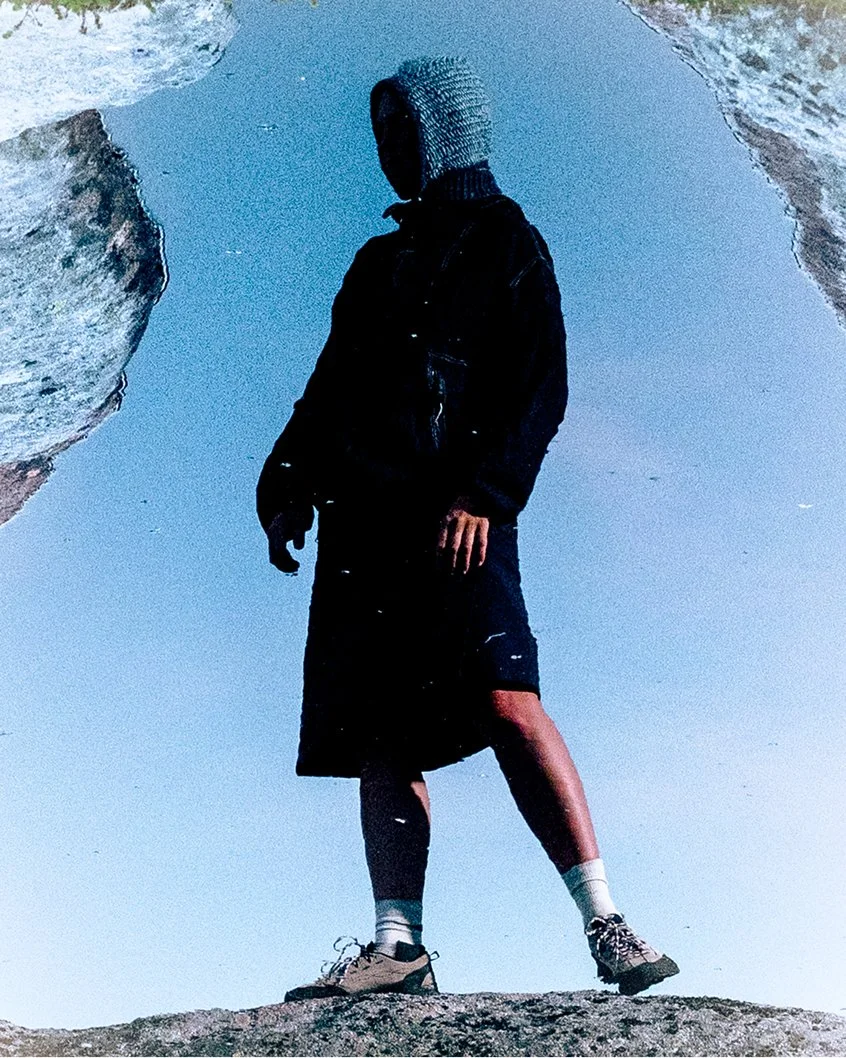 Person standing on rocks, wearing a hooded jacket, skirt, and a blue Clara Knitwear Brenna Hood, with their reflection showing in a water surface, creating an illusion of an upside-down reflection.