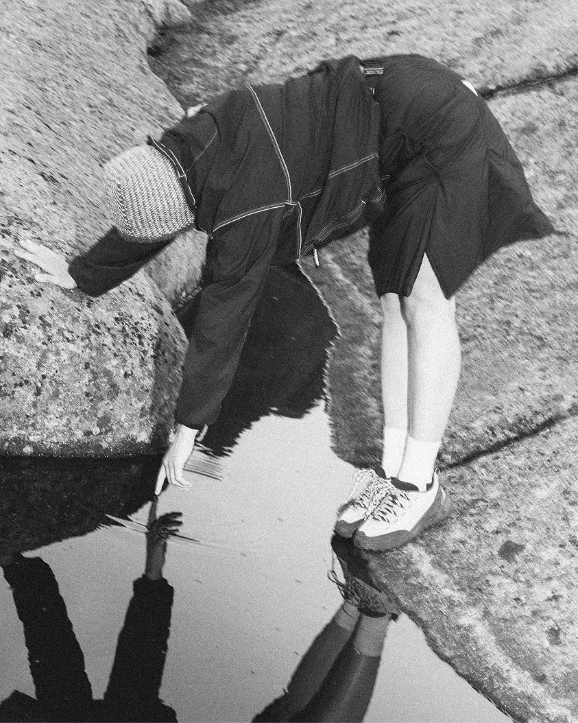 Person in a jacket, skirt, sneakers, and Clara Knitwear Brenna Hood crouching on rocks and reaching into water at the edge of still water.