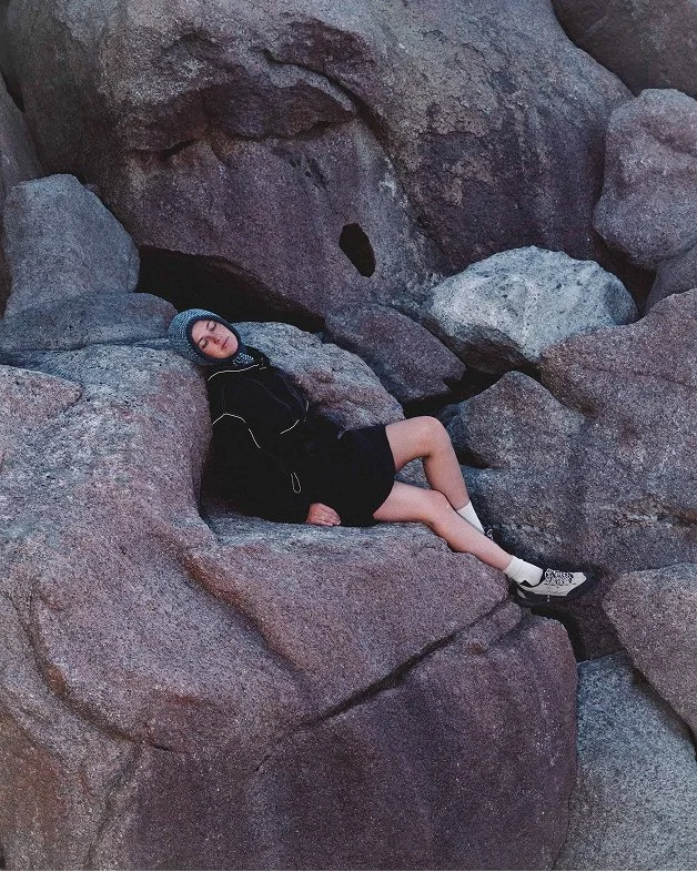 A person wearing a black jacket, black skirt, and a blue Clara Knitwear Brenna Hood, lying on large rock formations in a rocky outdoor setting.