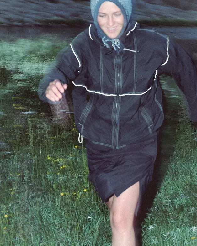 Person running outdoors on grassy area, wearing a black jacket, a skirt, and a blue Clara Knitwear Brenna Hood, with a smiling expression.