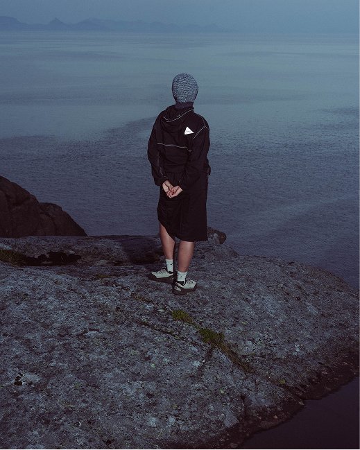 Person in black jacket and skirt standing on a rocky shoreline, looking out over calm water during dusk or dawn.