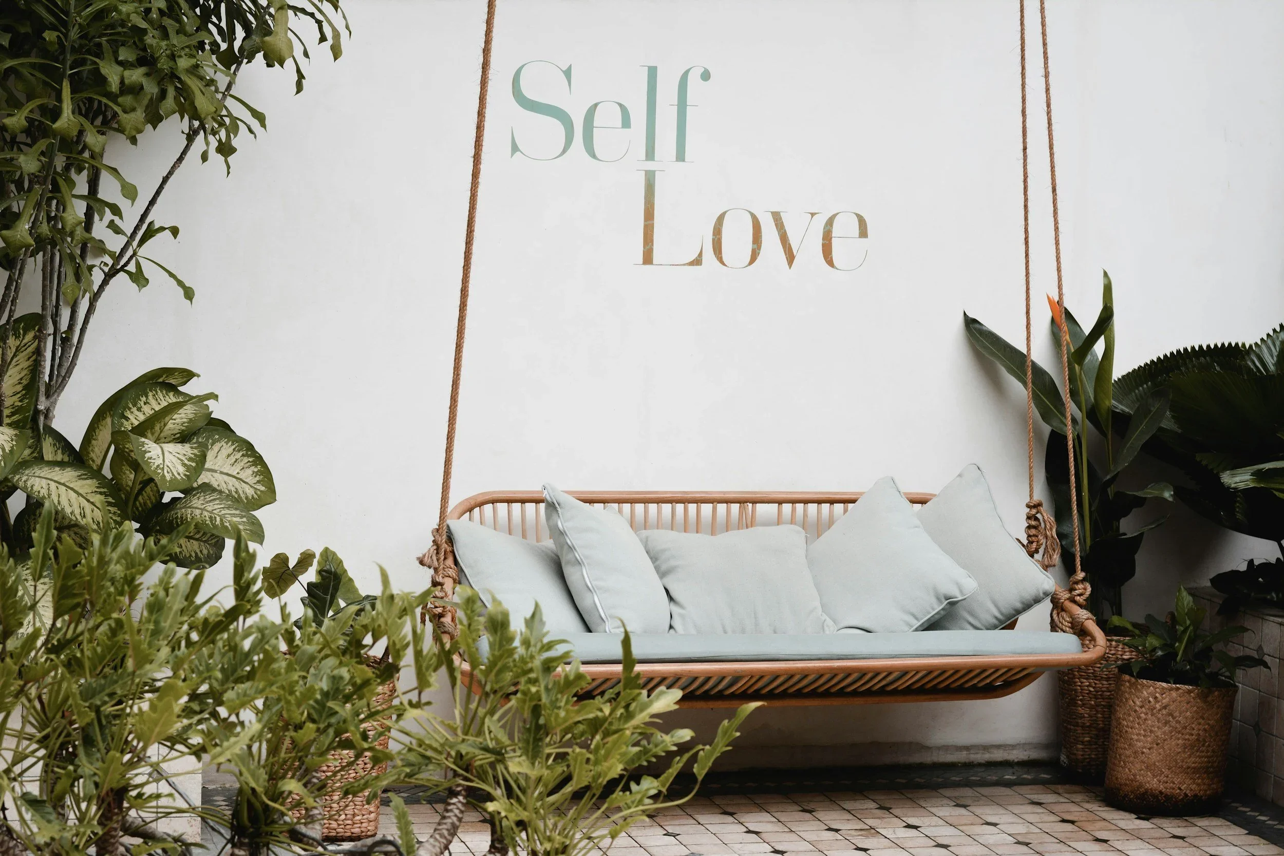 A peaceful patio with a hanging rattan swing, surrounded by plants and a wall reading 'Self Love,' representing the self-care and radical self-love practices taught by trauma therapist Mariah J. Zur.