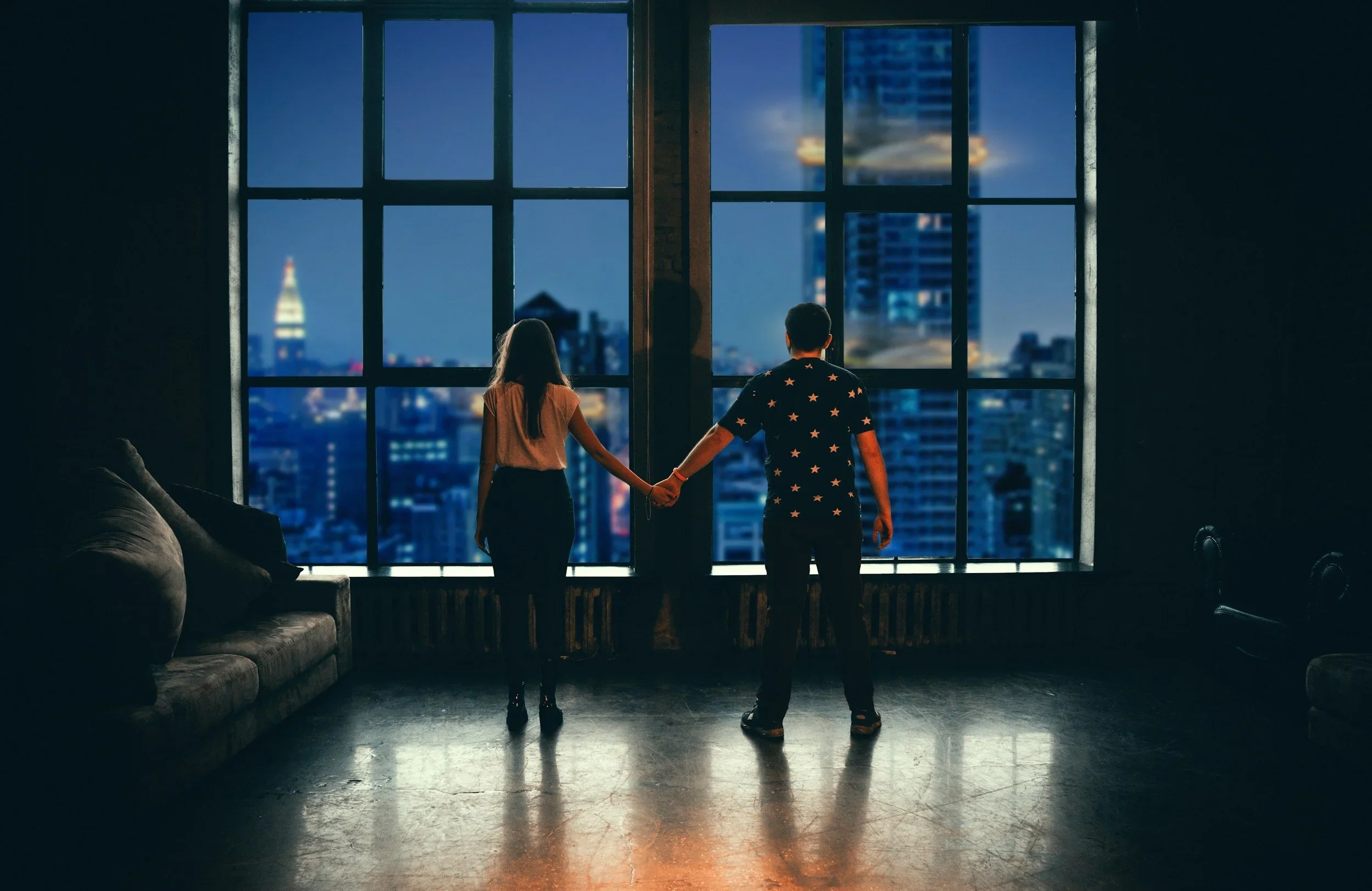 A couple holding hands and looking out a large window at a nighttime city skyline, representing relationship connection and healing.