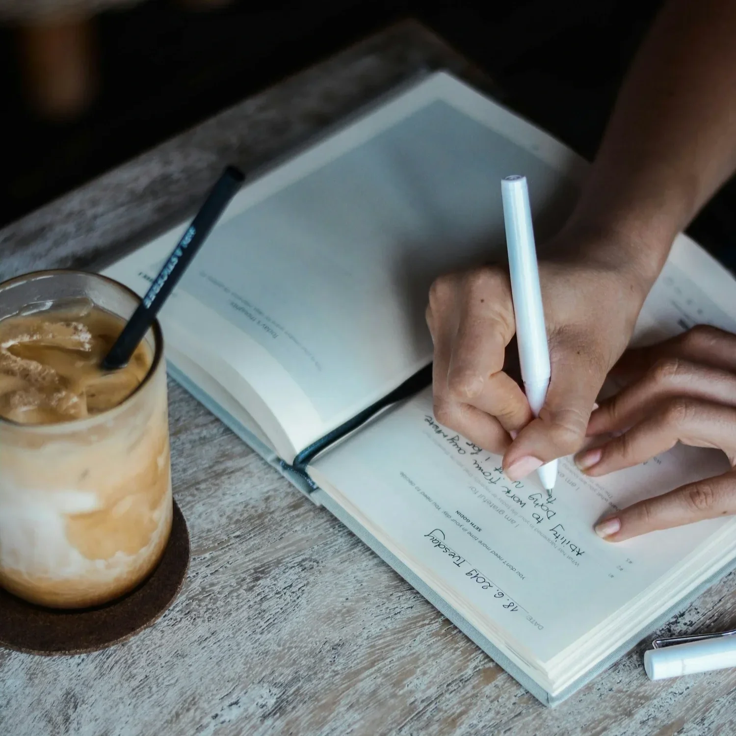 Person writing in a gratitude journal next to an iced coffee. By therapist Mariah J. Zur.