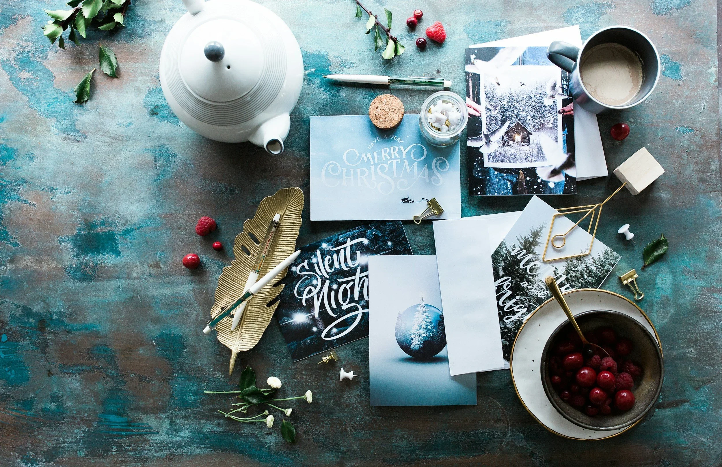 Flat lay of holiday greeting cards, a teapot, and berries on a desk, representing the busy preparation of the holiday season. By therapist Mariah J. Zur.