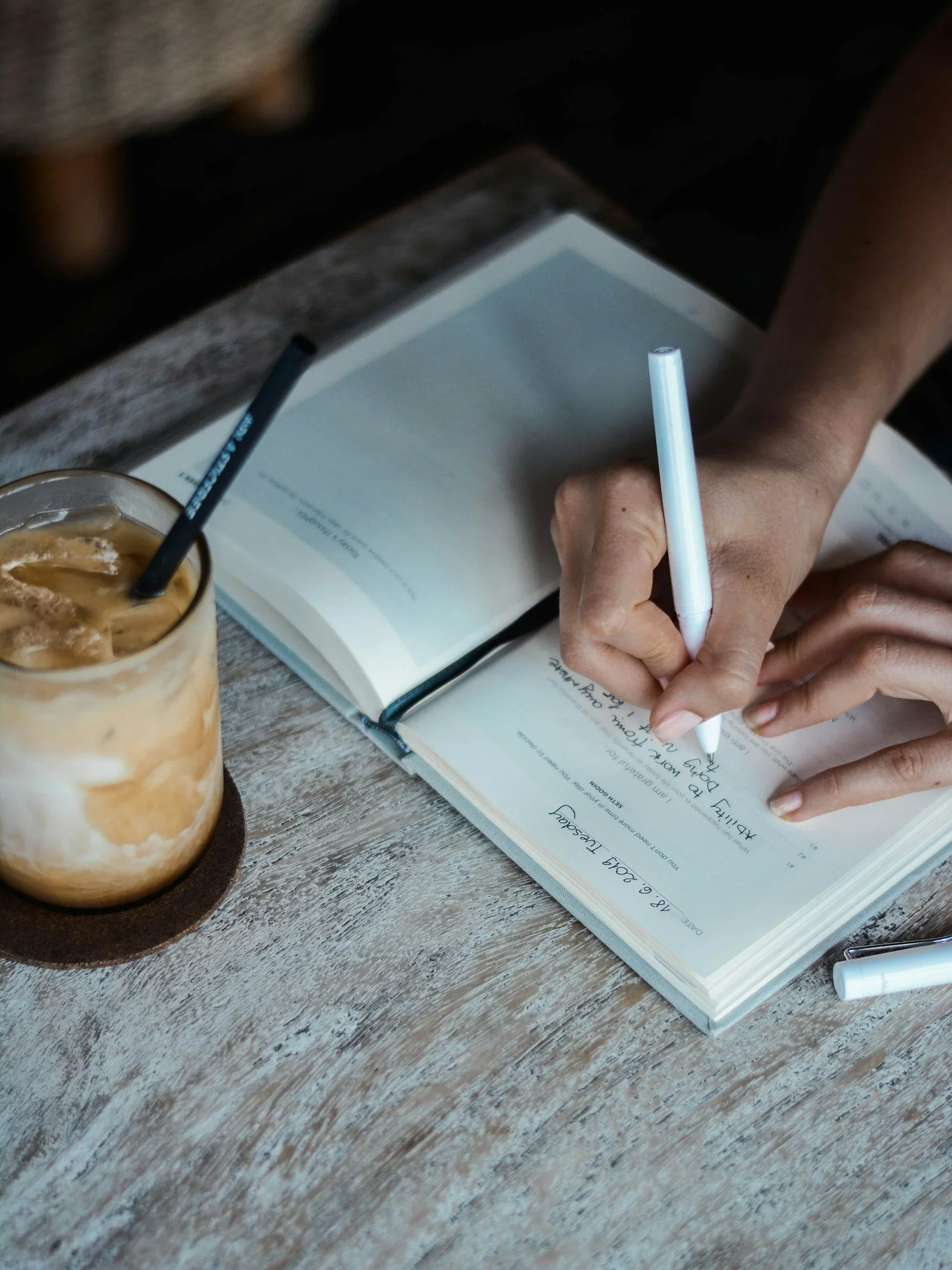 A person writing in a recovery workbook next to an iced coffee, practicing the daily integration of healing strategies taught through psychological trauma books and therapy intensives in Raleigh, NC.