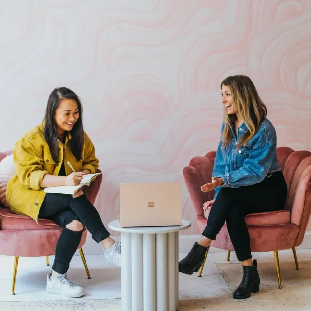 Two female professionals sitting in pink velvet chairs in a modern office, engaged in an upbeat discussion with a laptop and notebook, representing a collaborative trauma therapy intensive session with Mariah J. Zur.
