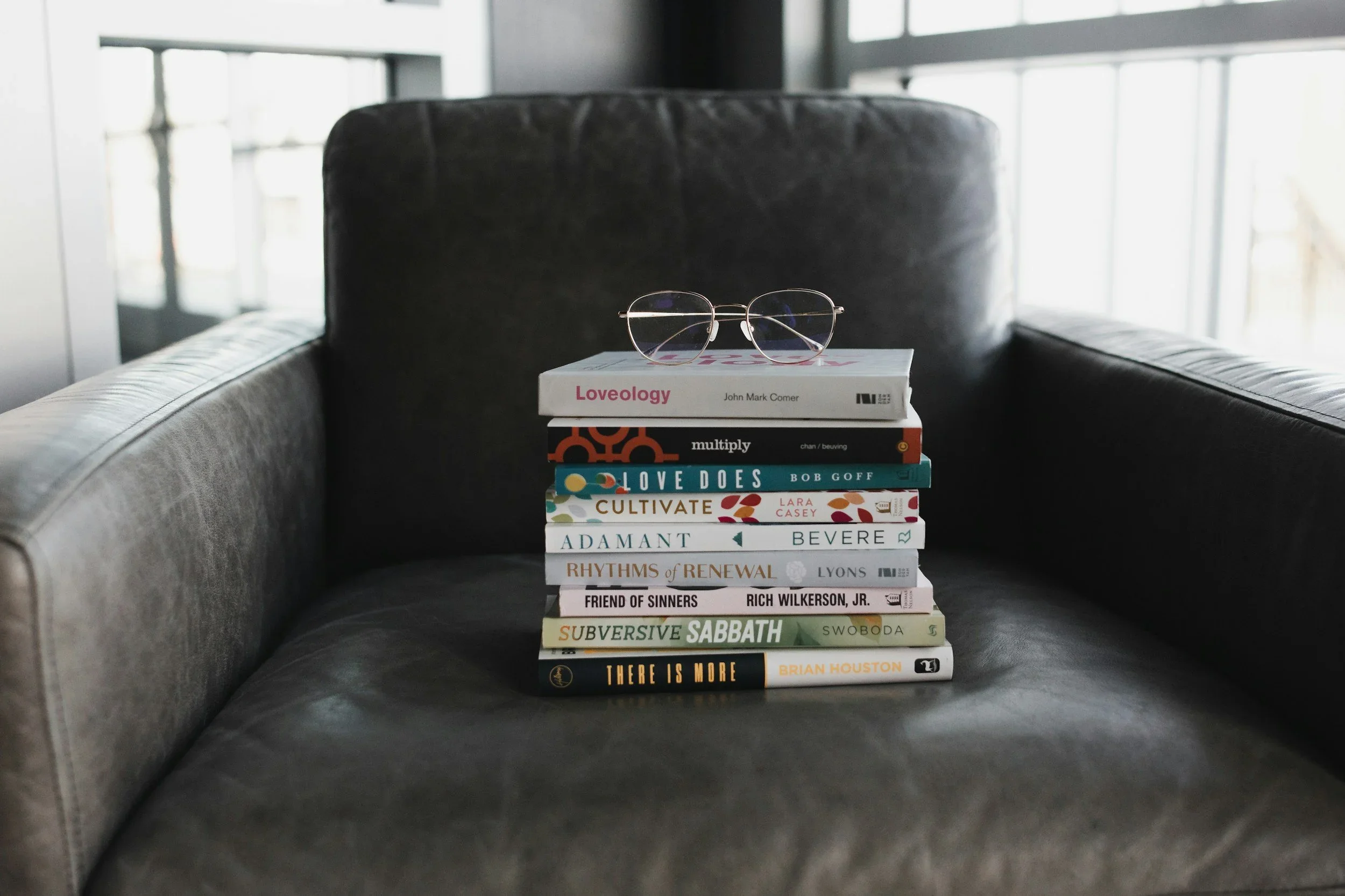 A stack of nine specific Christian non-fiction book spines on a grey leather armchair seat, listing all titles including Loveology, Multiply, Love Does, Cultivate, Adamant, Rhythms of Renewal, Friend of Sinners, Subversive Sabbath, and There is More.