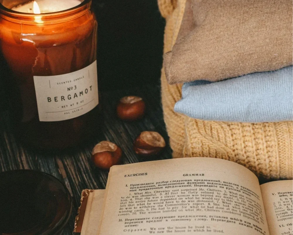 A cozy setup with a lit bergamot candle, a stack of sweaters, and an open book. PA therapist Mariah Zur encourages grounding activities like reading and creating a calm environment for mental wellness.