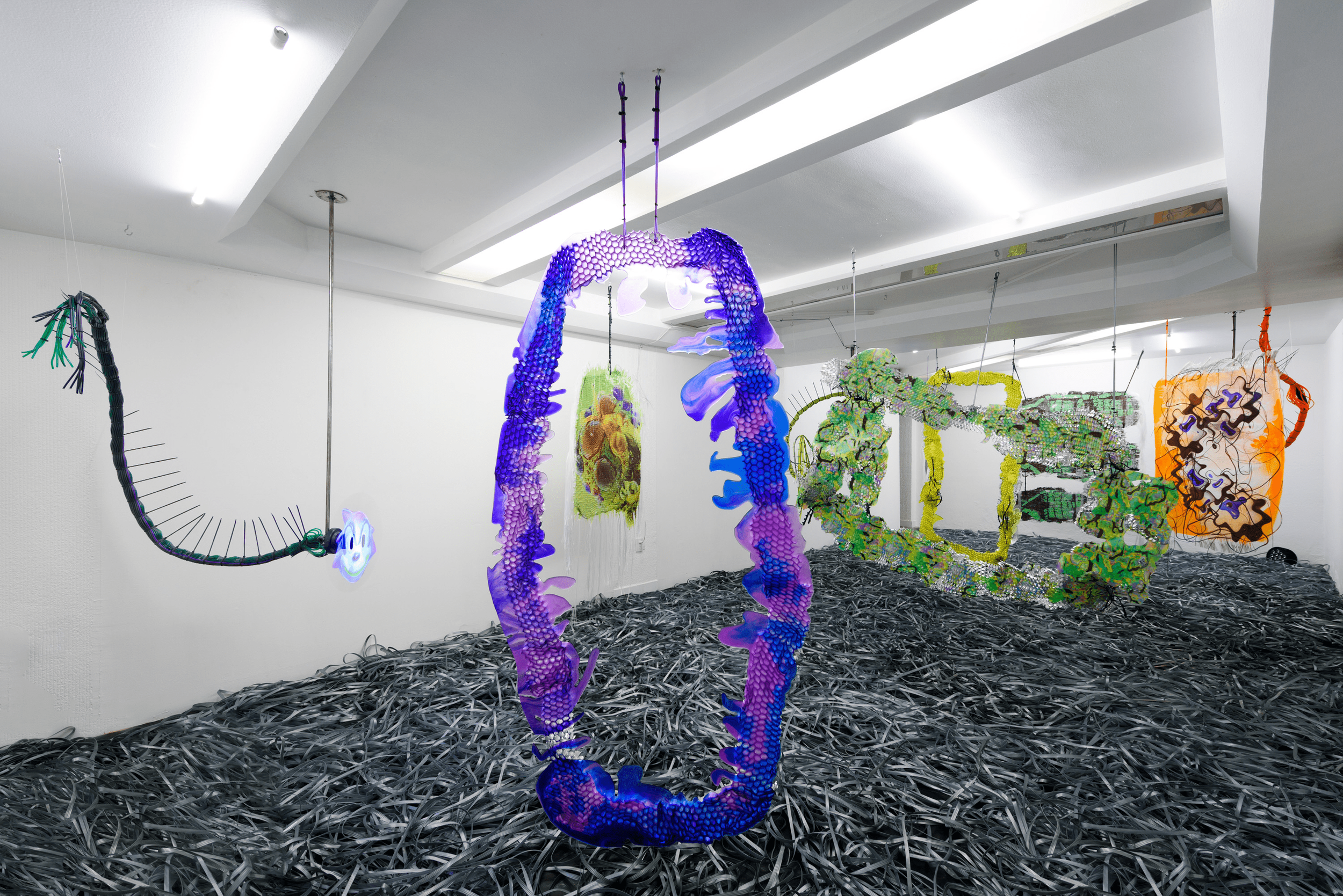 Contemporary art installation in a gallery, featuring large, colorful, abstract sculptures suspended from the ceiling and a floor covered in black, tangled materials.