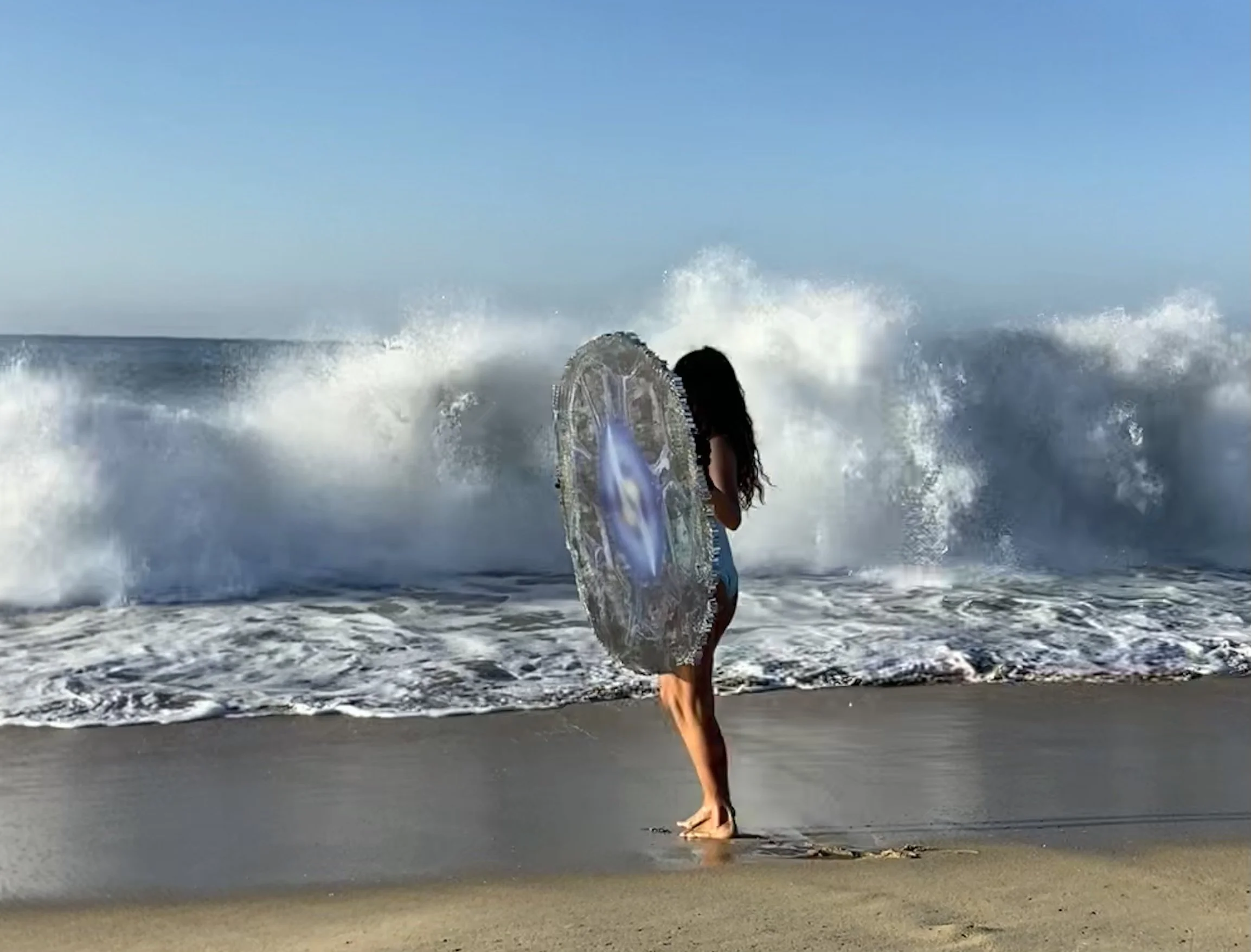 Woman standing on the beach with a surfboard, facing the ocean with waves crashing in the background.