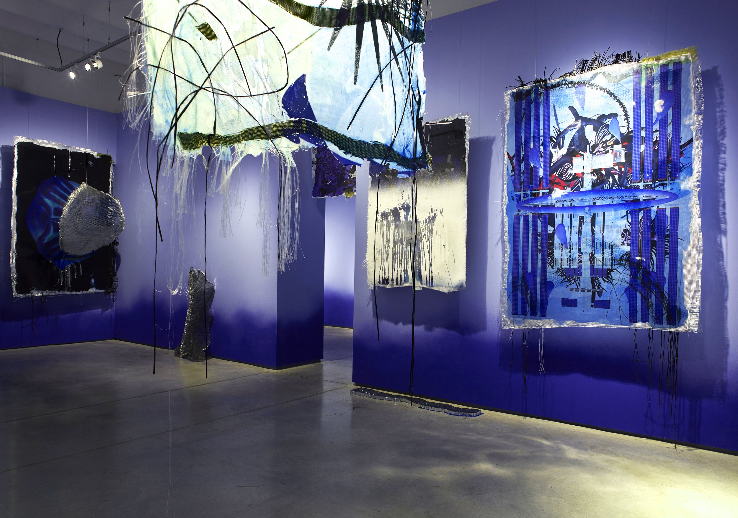 Art installation featuring mixed media textiles and fabric art pieces on walls and hanging from ceiling in an art gallery with purple walls and concrete floor.