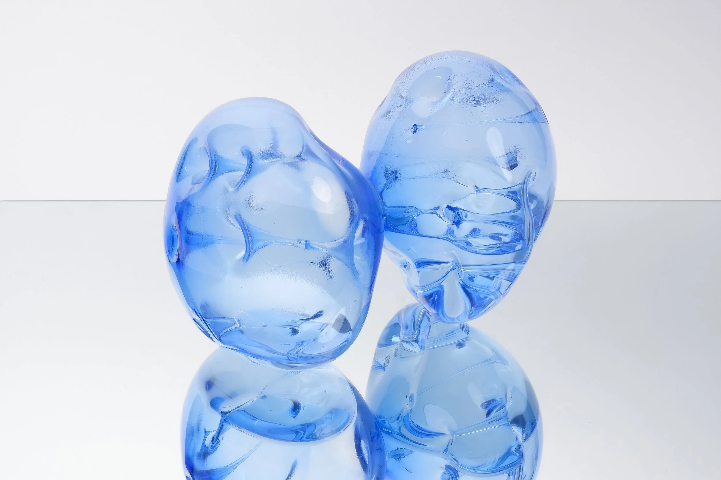 Two abstract blue glass sculptures on a reflective surface.