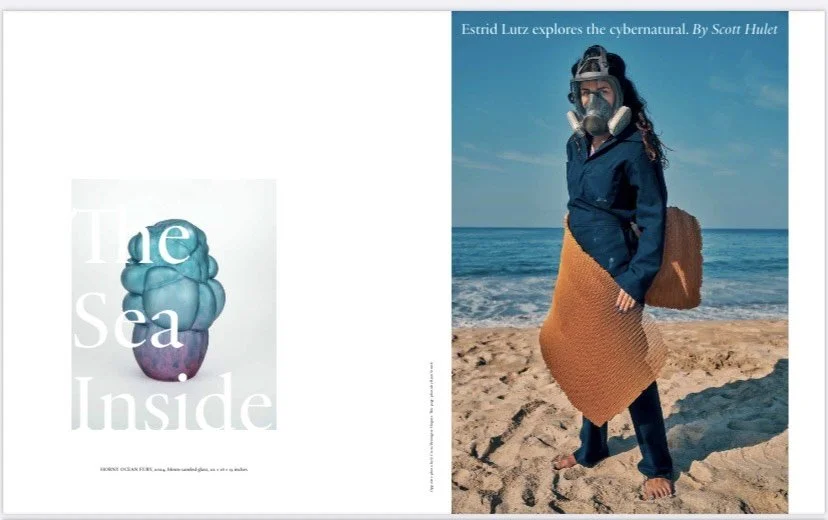 An open magazine displaying two pages. The left page has a title "The Sea Inside" with an abstract sculpture that resembles a cloud or ocean wave. The right page shows a woman on a beach wearing a mask and snorkel, a coat, a brown skirt, and a large shoulder bag, with an ocean and cloudy sky in the background.