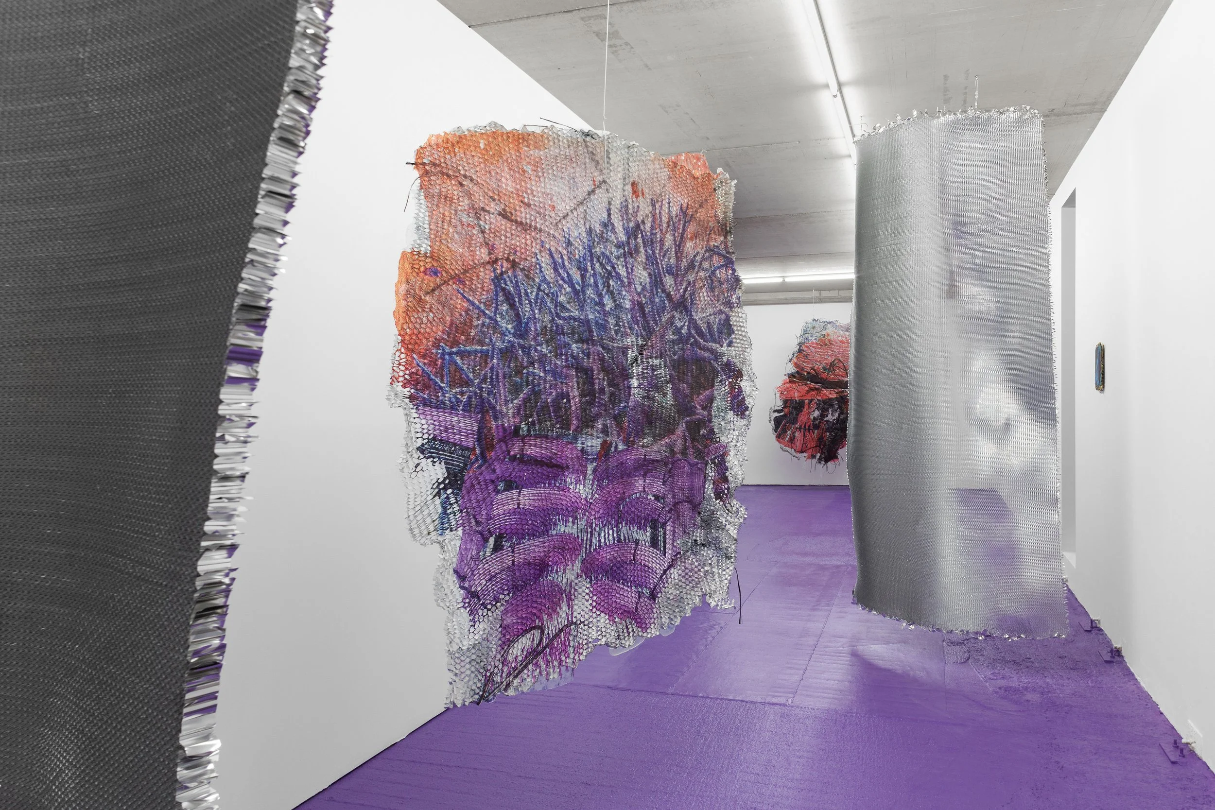 Contemporary art installation featuring fabric, mesh, or textured materials hanging in an exhibition space with white walls and purple floors.