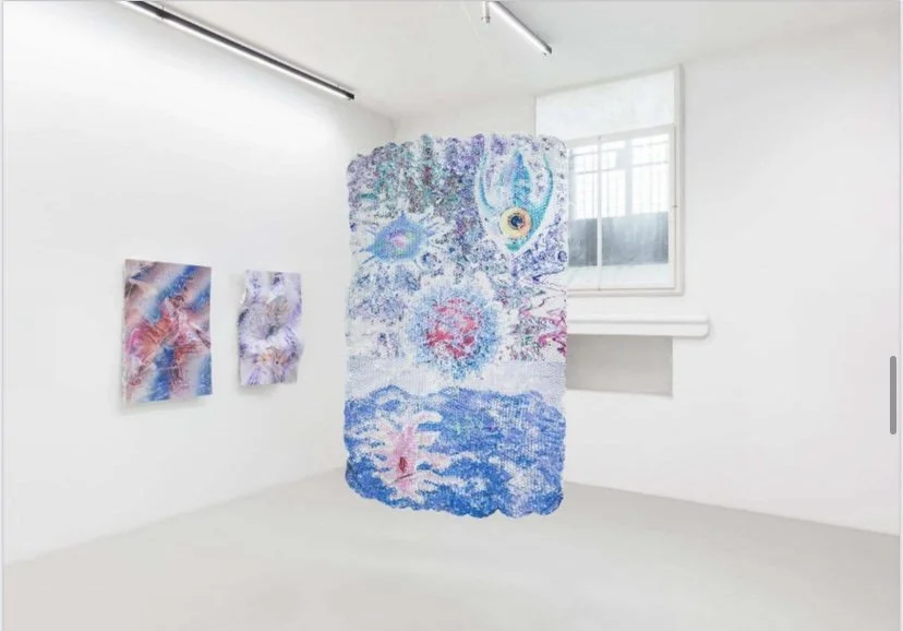 Contemporary art exhibit in a white gallery featuring three colorful abstract paintings and a hanging textile piece with vibrant patterns.