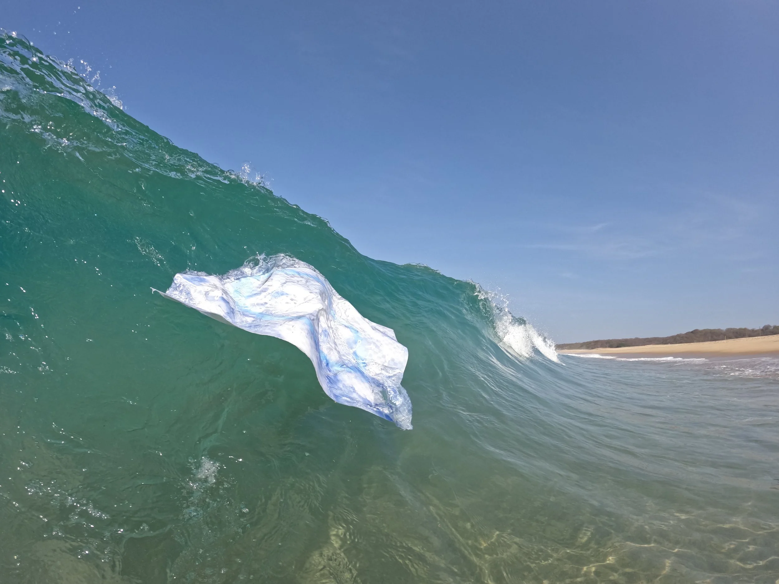 Image of a clear ocean wave with a piece of plastic floating inside it, near a sandy beach under a blue sky.