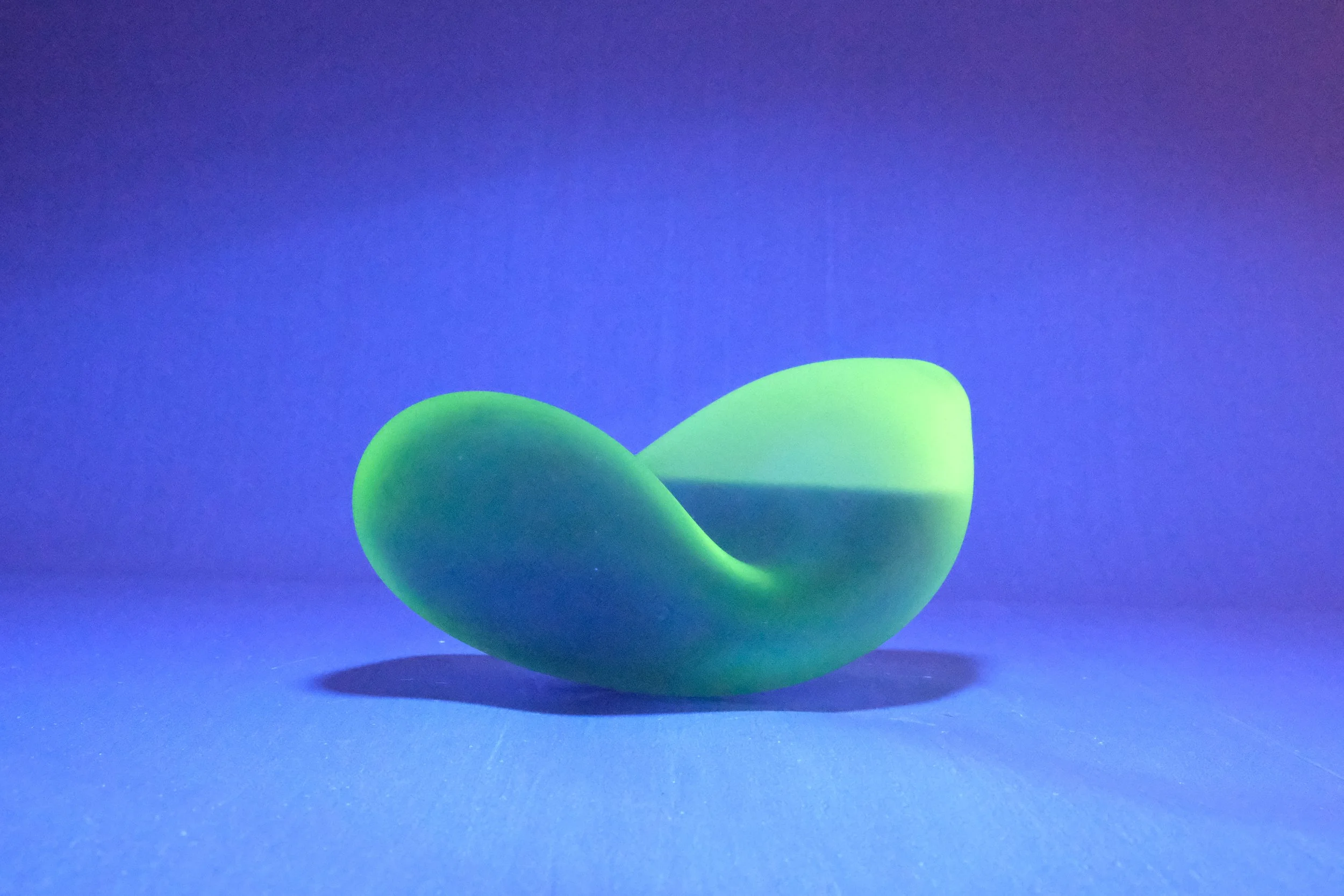 Green abstract sculpture with smooth, curved form on a blue background casting a shadow.