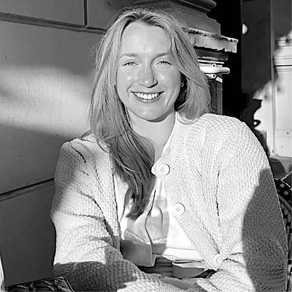 Black and white photo of a woman smiling, sitting indoors with books and objects behind her.