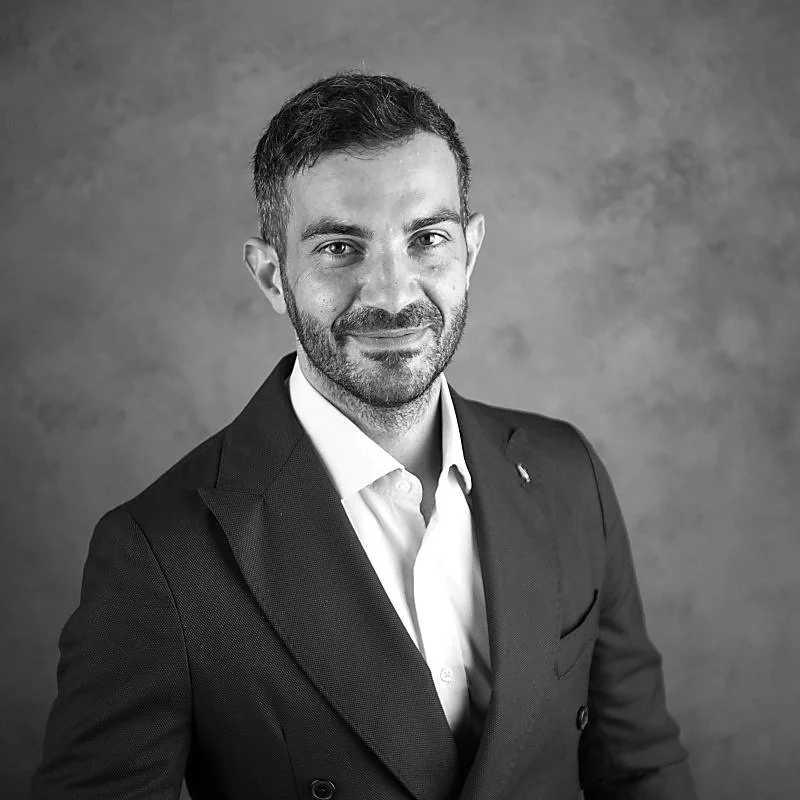 A professional man with short dark hair, beard, and mustache, wearing a dark blazer over a white shirt, smiling at the camera against a plain background.