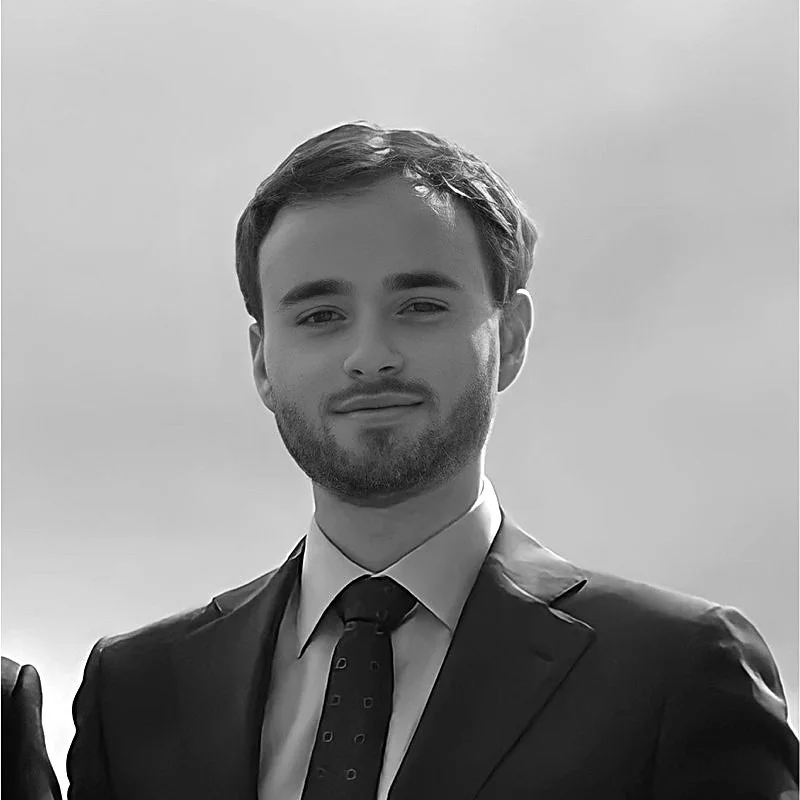 A young man with short hair, beard, wearing a suit and tie, standing outdoors with a cloudy sky in the background.