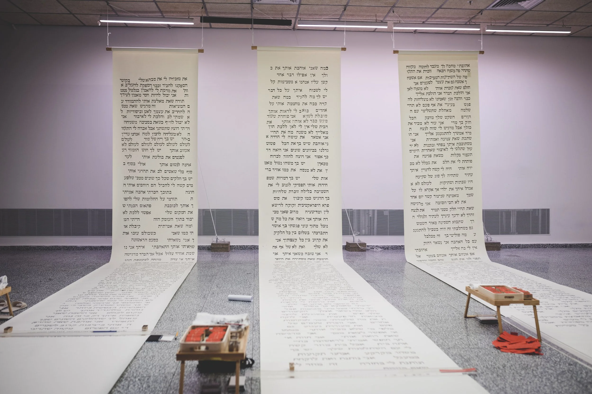 Three large vertical banners hanging from the ceiling with Hebrew text printed on them, set in an indoor space with a gray floor and white walls.