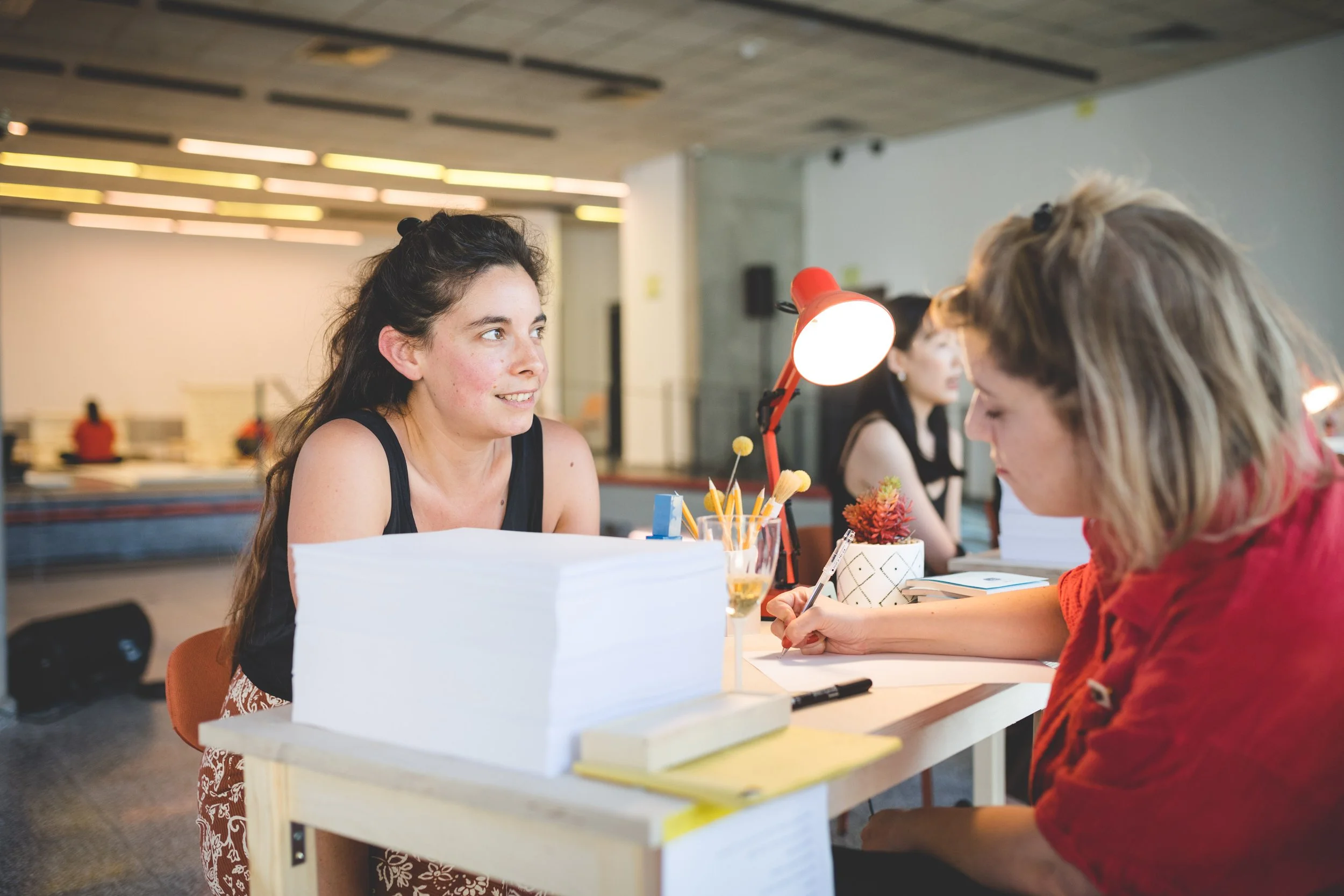 A woman sitting at a reception desk talking to a receptionist in a modern indoor setting, with other people in the background.