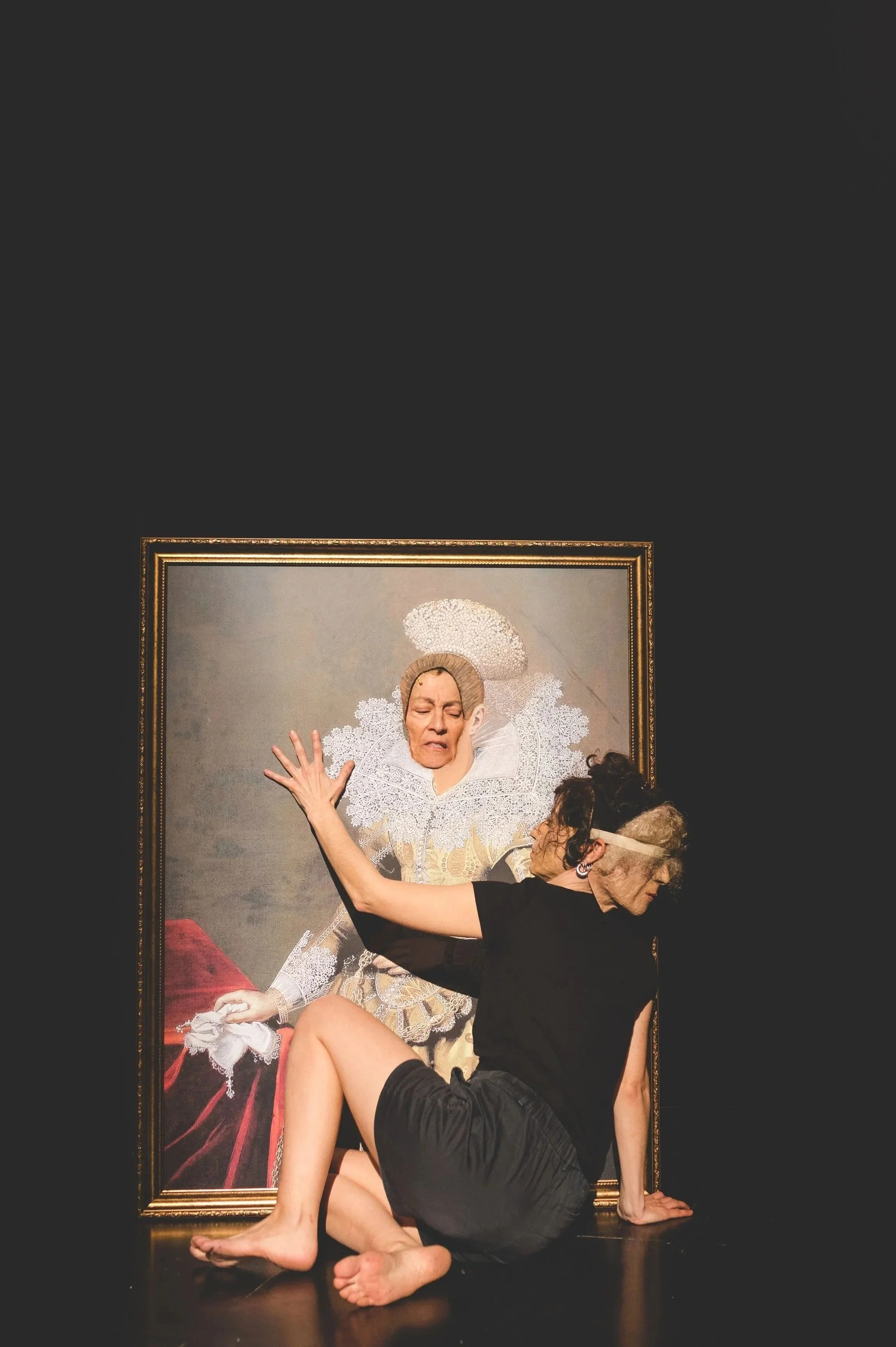 A woman with gray hair and glasses posing in front of a portrait of a woman in historical clothing, wearing a lace collar and headdress, with her hand raised high.