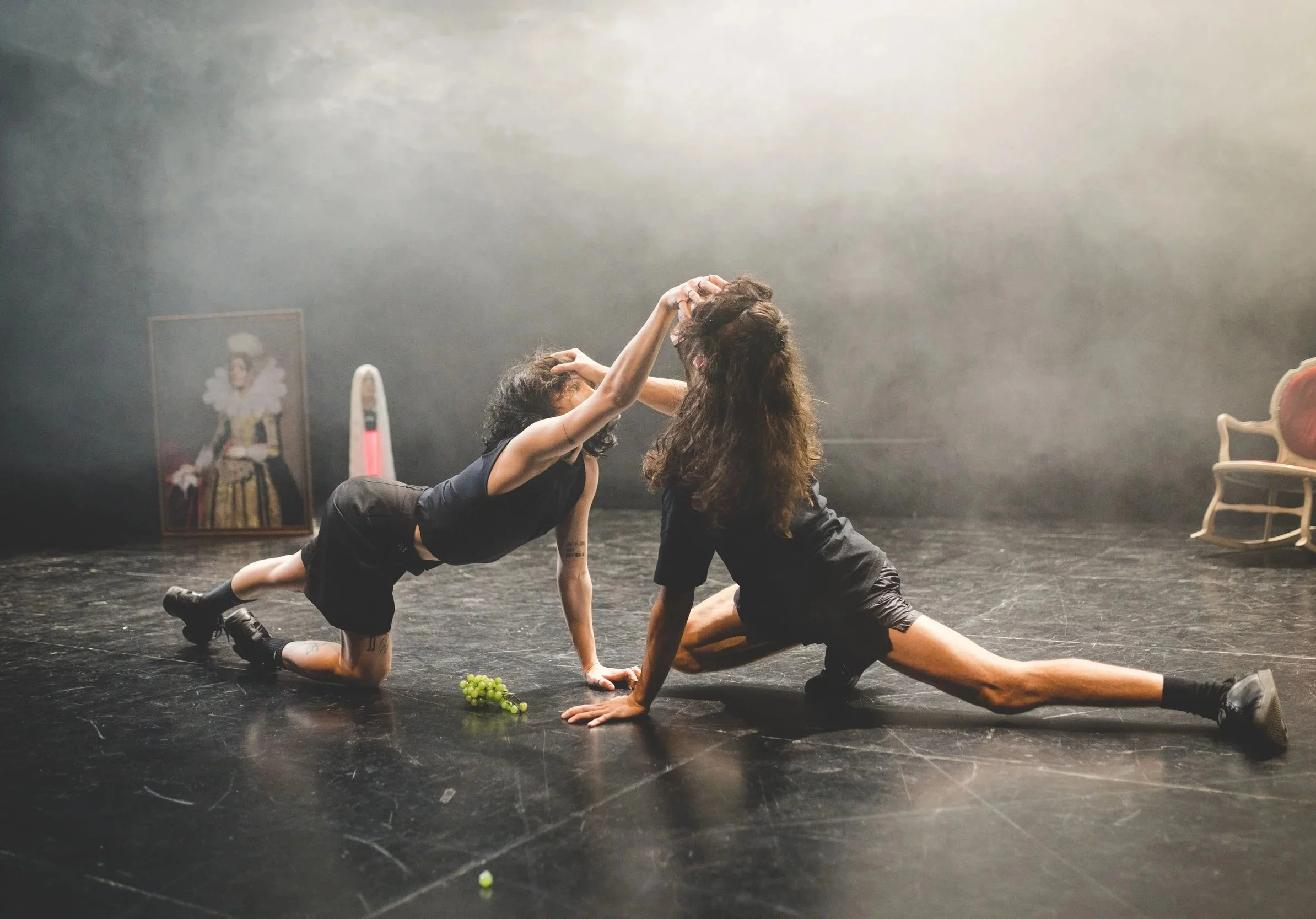 Two women in black clothing performing a theatrical dance or re-enactment on a black stage, one on all fours with one leg outstretched, the other on her knees leaning forward with arms raised. A grape cluster is on the floor nearby. Background includ