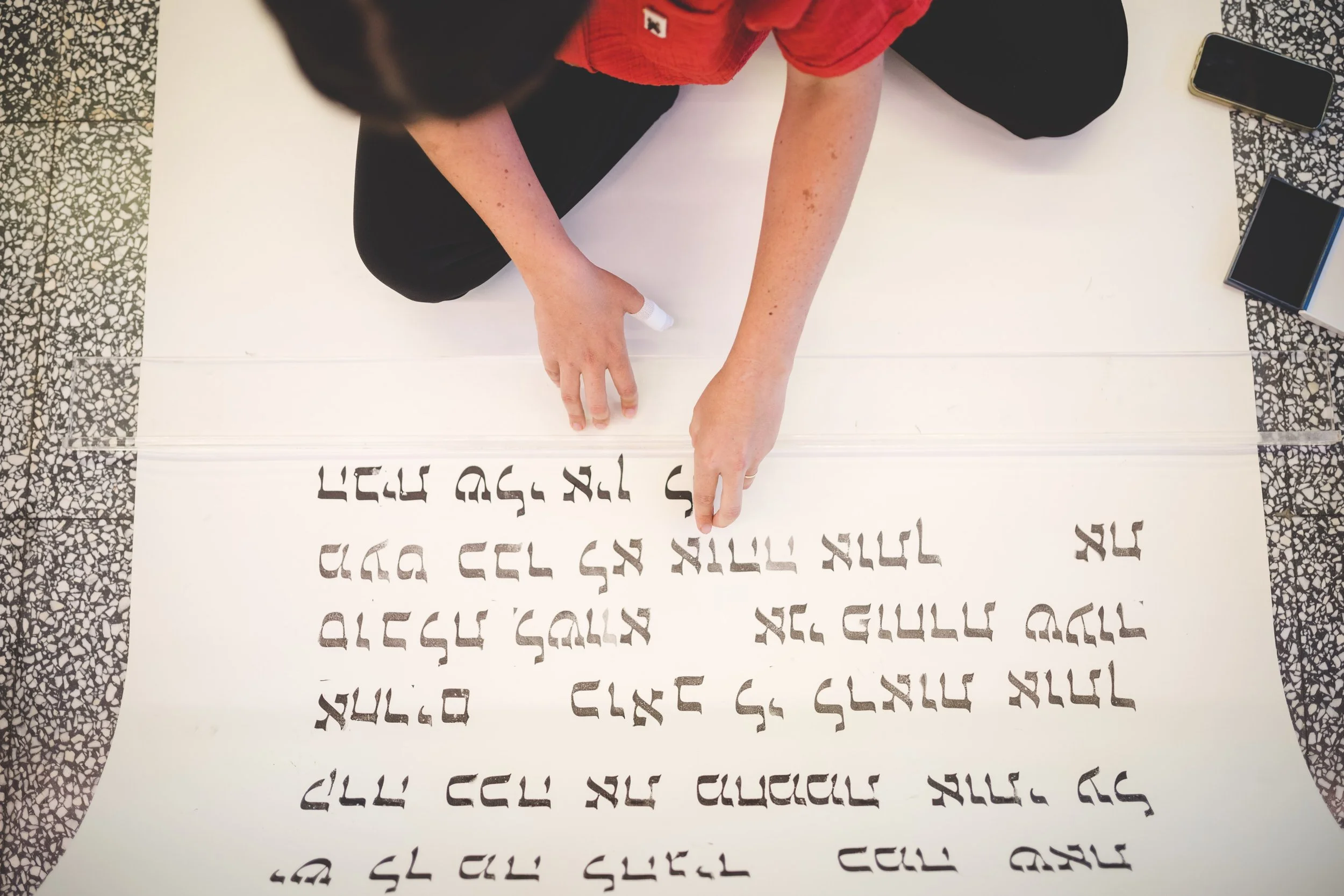 A person sitting on the floor writing or drawing on a large white sheet of paper with a black marker. The paper has various lines of text or symbols written in black ink. The person's head is not visible, but they are wearing a red shirt and black pa