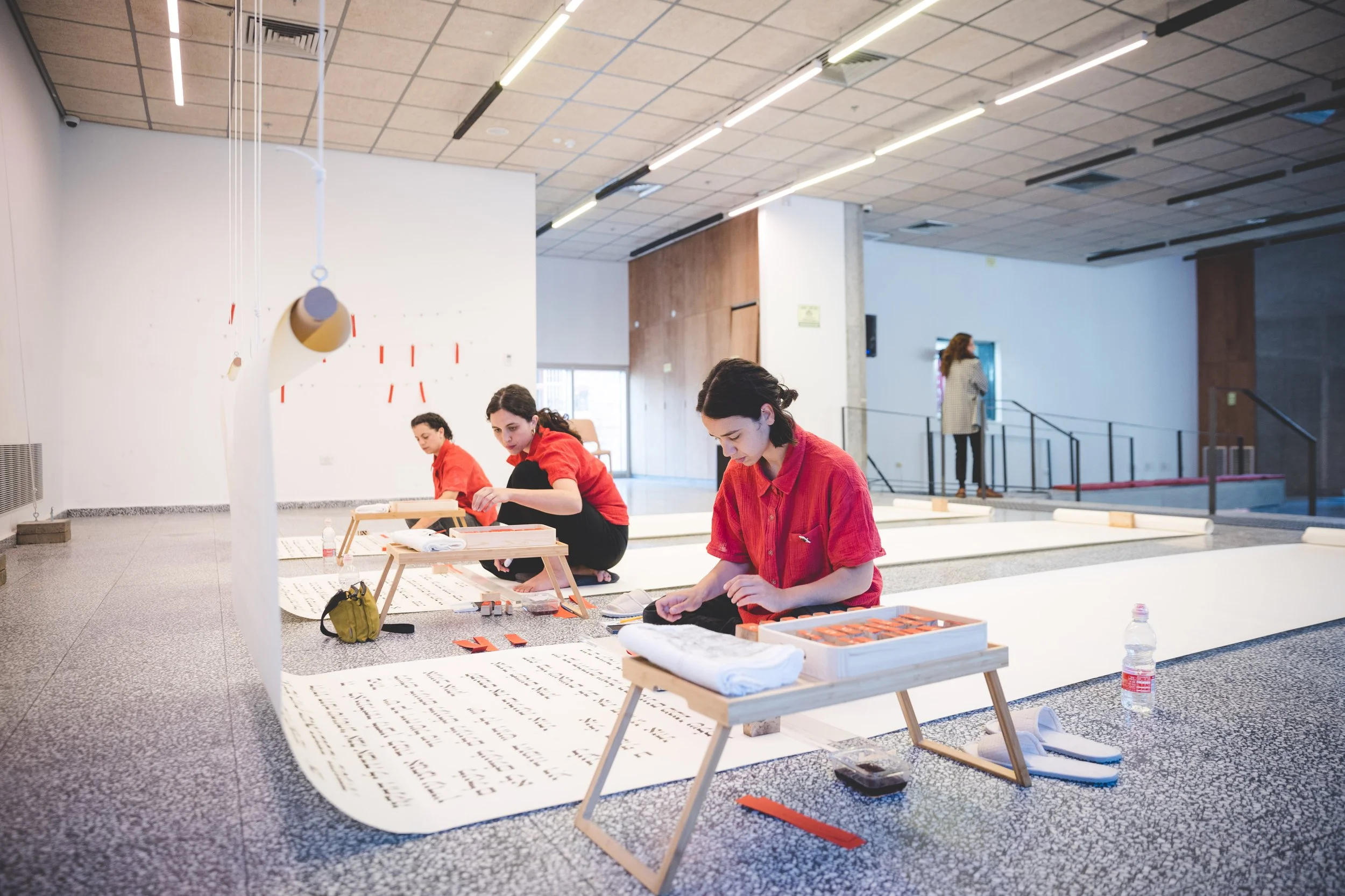 Three women in red shirts working on calligraphy with large sheets of paper on the floor in an indoor art space, with another woman standing in the background.