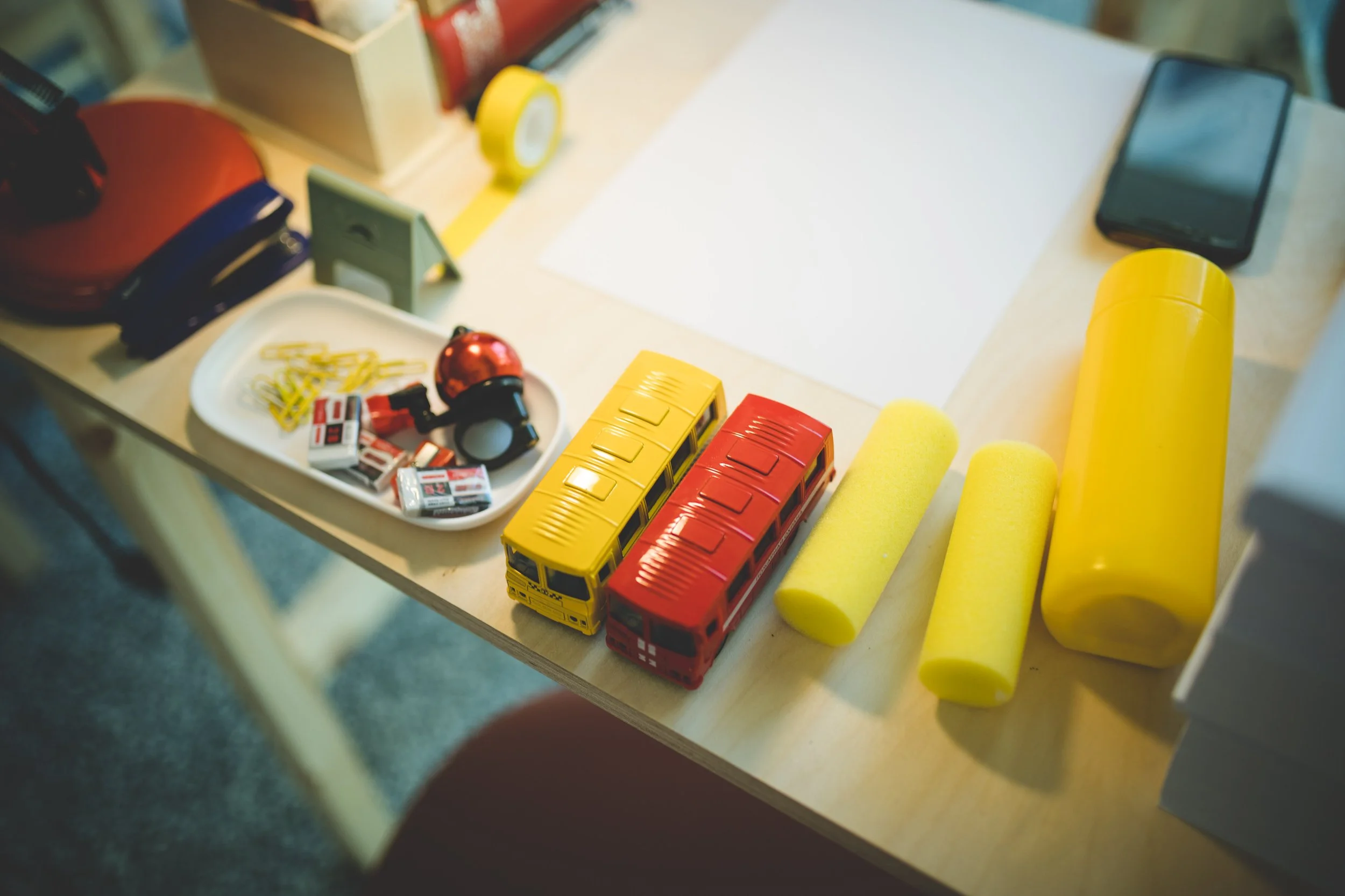 Children's toys and supplies on a desk, including two yellow and one red toy buses, two yellow foam rollers, a yellow water bottle, a black smartphone, a white tray with small toys, and a red storage container.