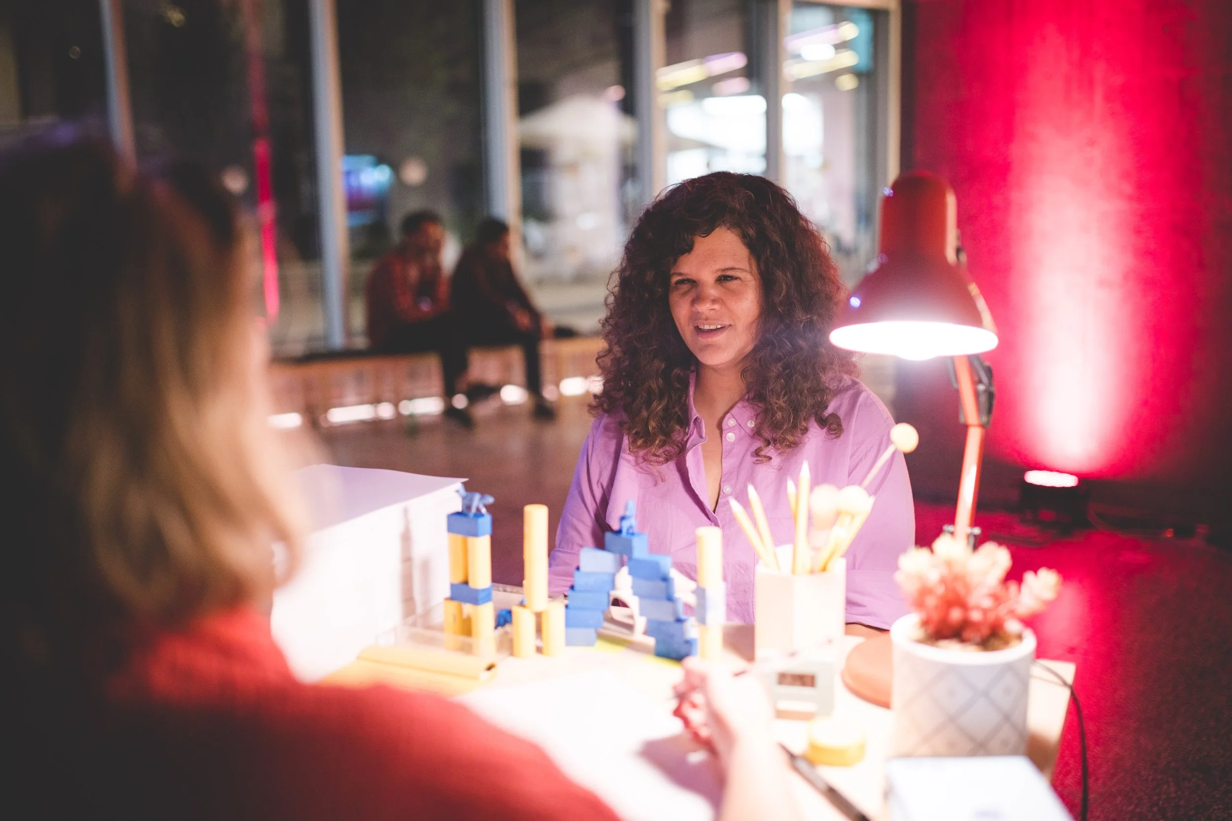 A woman with curly hair sitting at a desk and talking to another person in a cafe. The desk has a science kit with building blocks, test tubes, and a lamp. There are other people in the background sitting by the window.