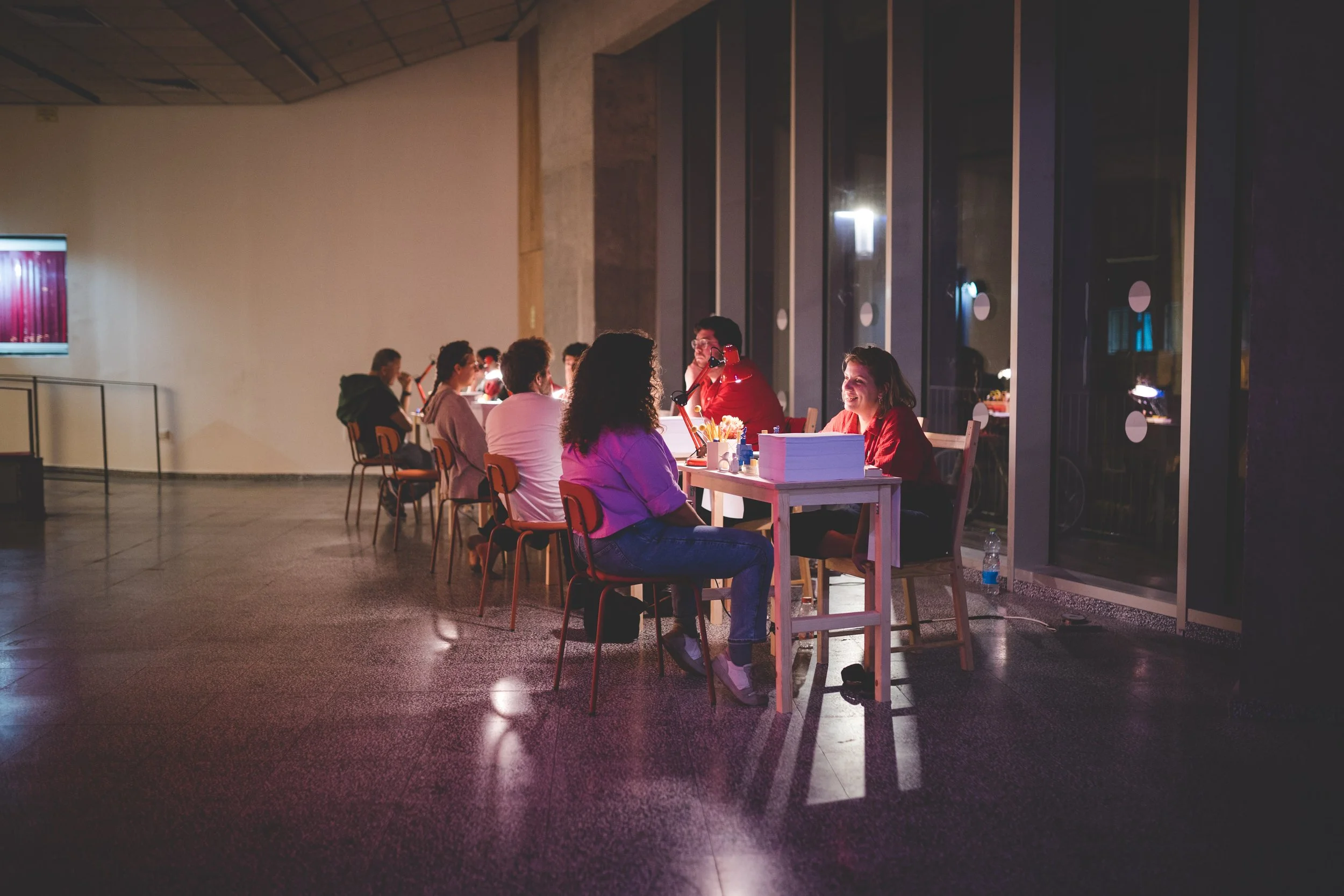 A group of people seated at tables in a modern indoor space, engaging in conversation and enjoying drinks, illuminated by soft lighting.