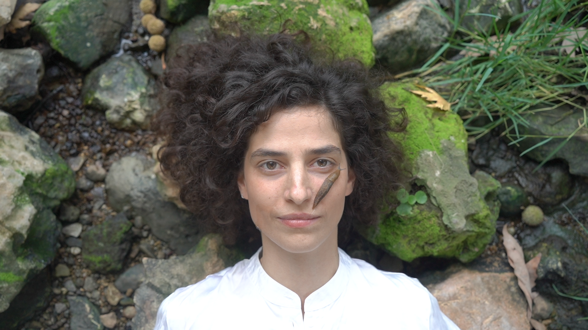 A woman with curly dark hair lying on a bed of moss-covered rocks and greenery, with a small brown and gray snail on her right cheek.