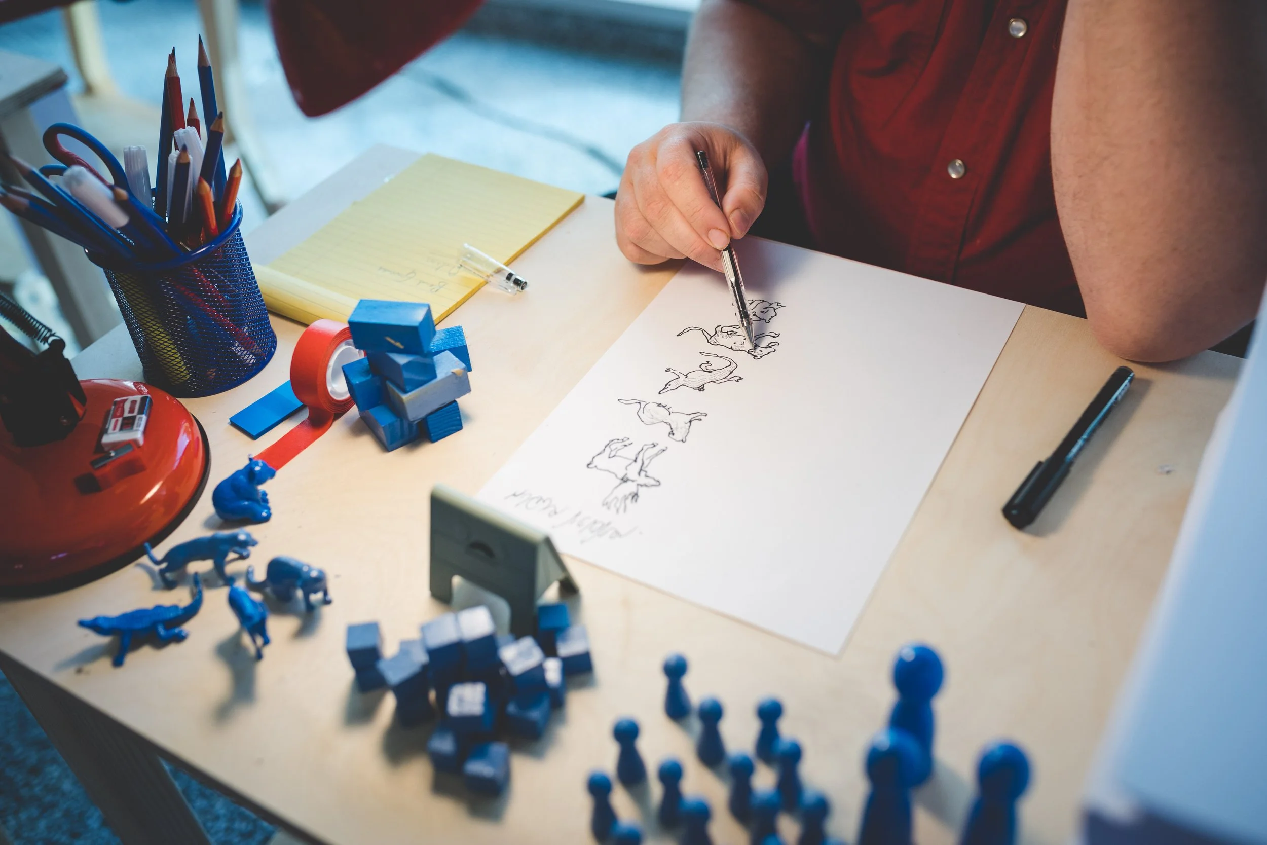 Person drawing animals like a horse, elephant, and dog on a sheet of paper at a desk filled with yellow notepad, pens, and blue toys.