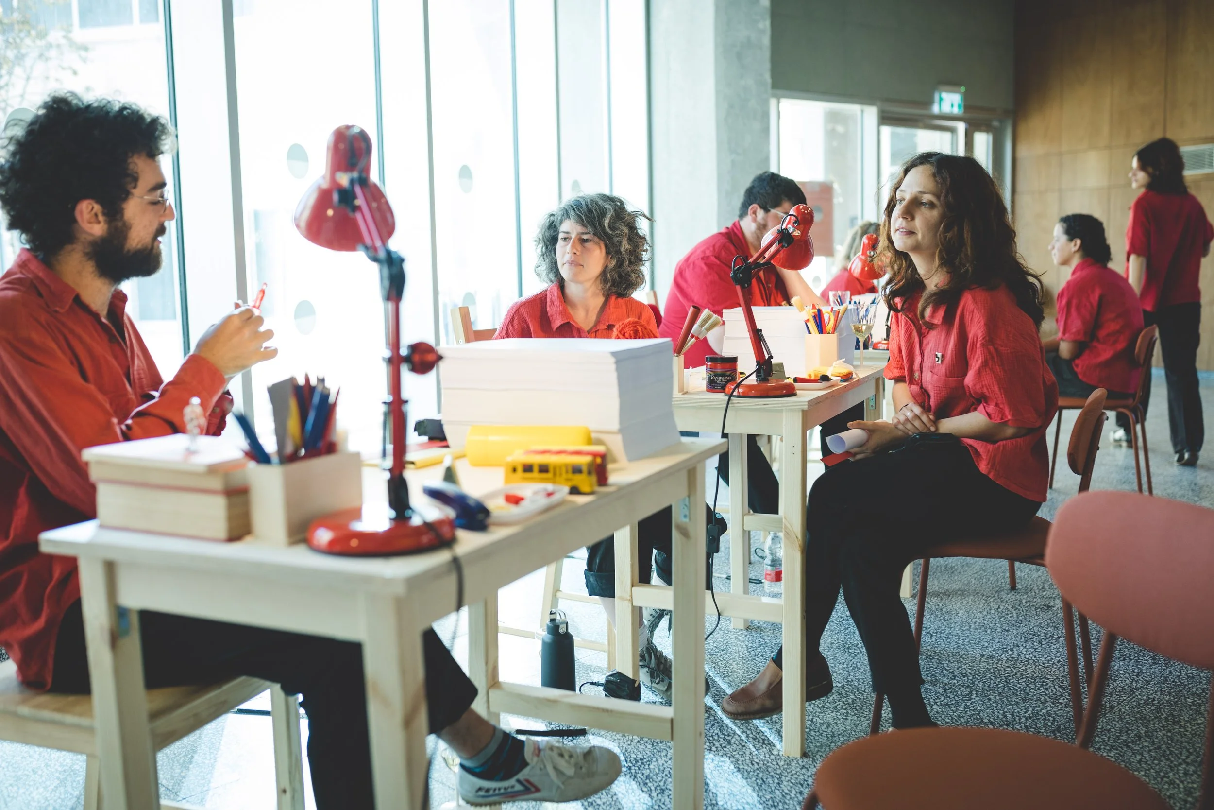 Group of people in red shirts working on arts and crafts at tables with lamps, surrounded by supplies in a bright room with large windows.