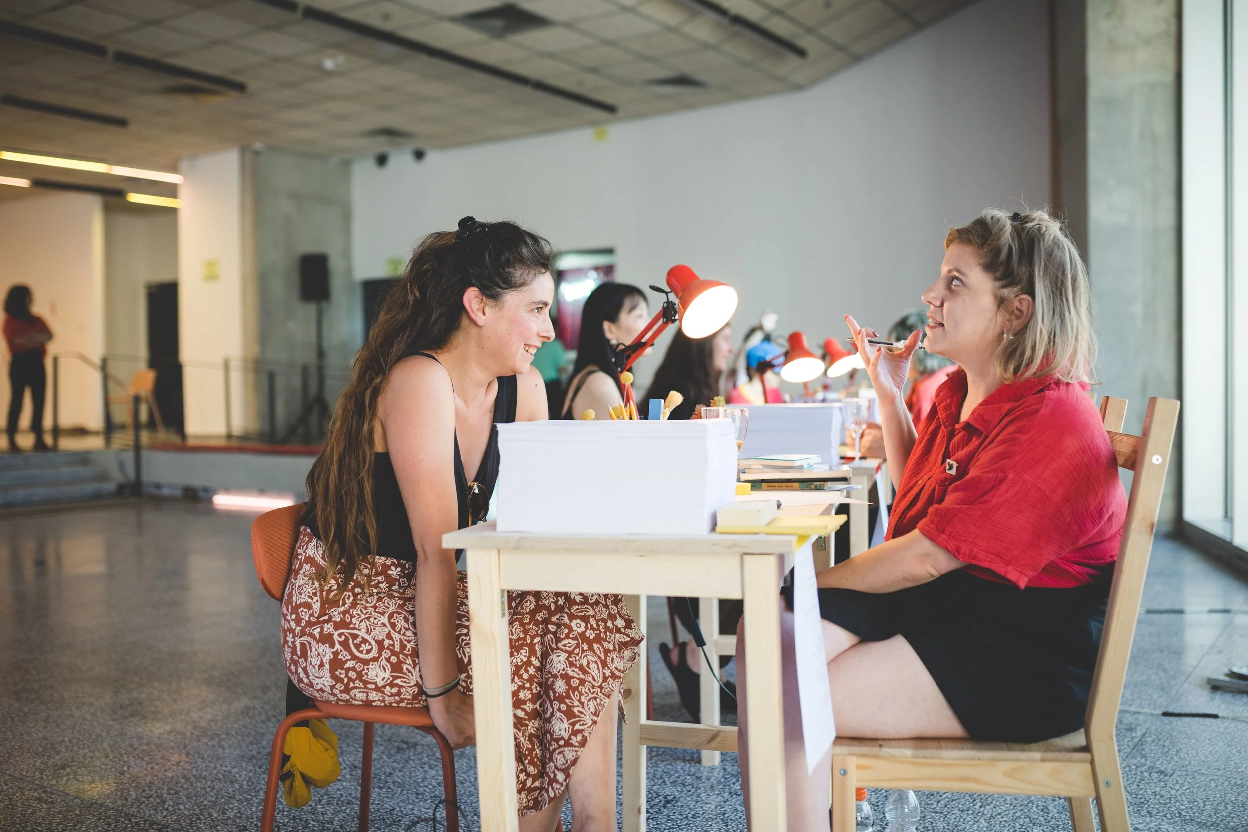 Two women having a conversation at a desk in a modern indoor setting, with other people working in the background.