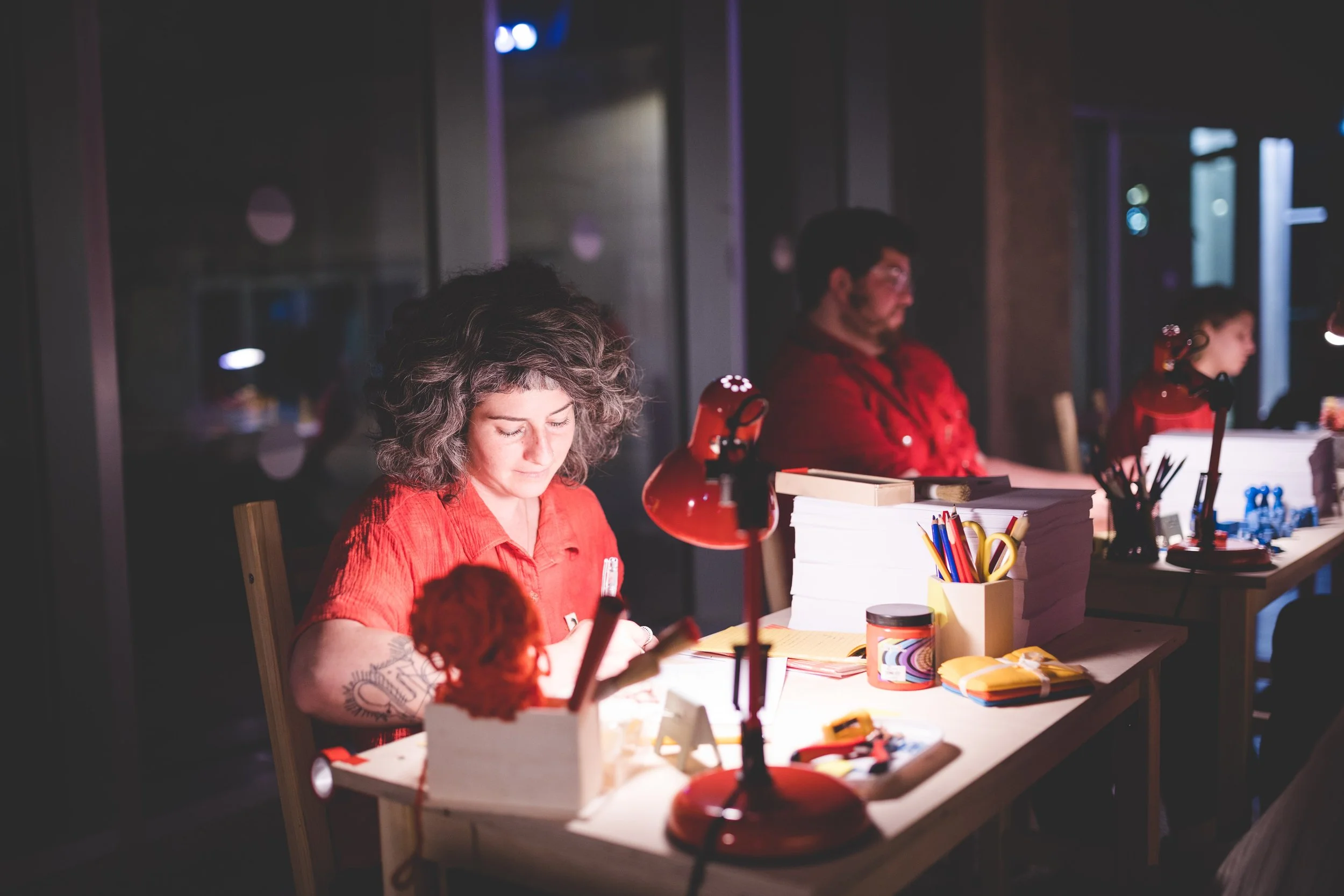 Three people sitting at a desk, working in a dimly lit room with lamps, pens, books, and office supplies on the desk.