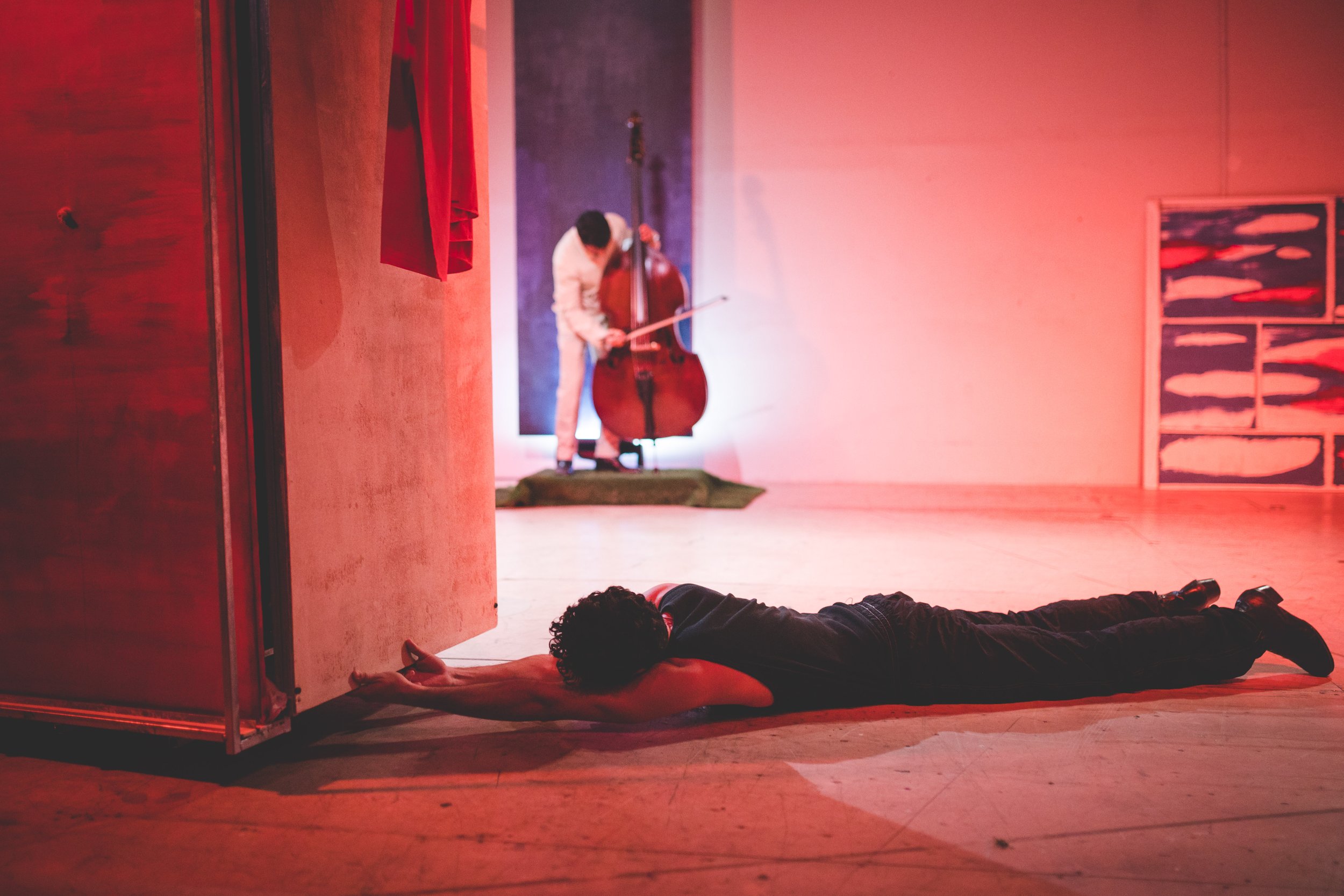 Person lying face down on the floor with arms extended forward, in front of a partially open door, with a musician playing a double bass on stage in the background.