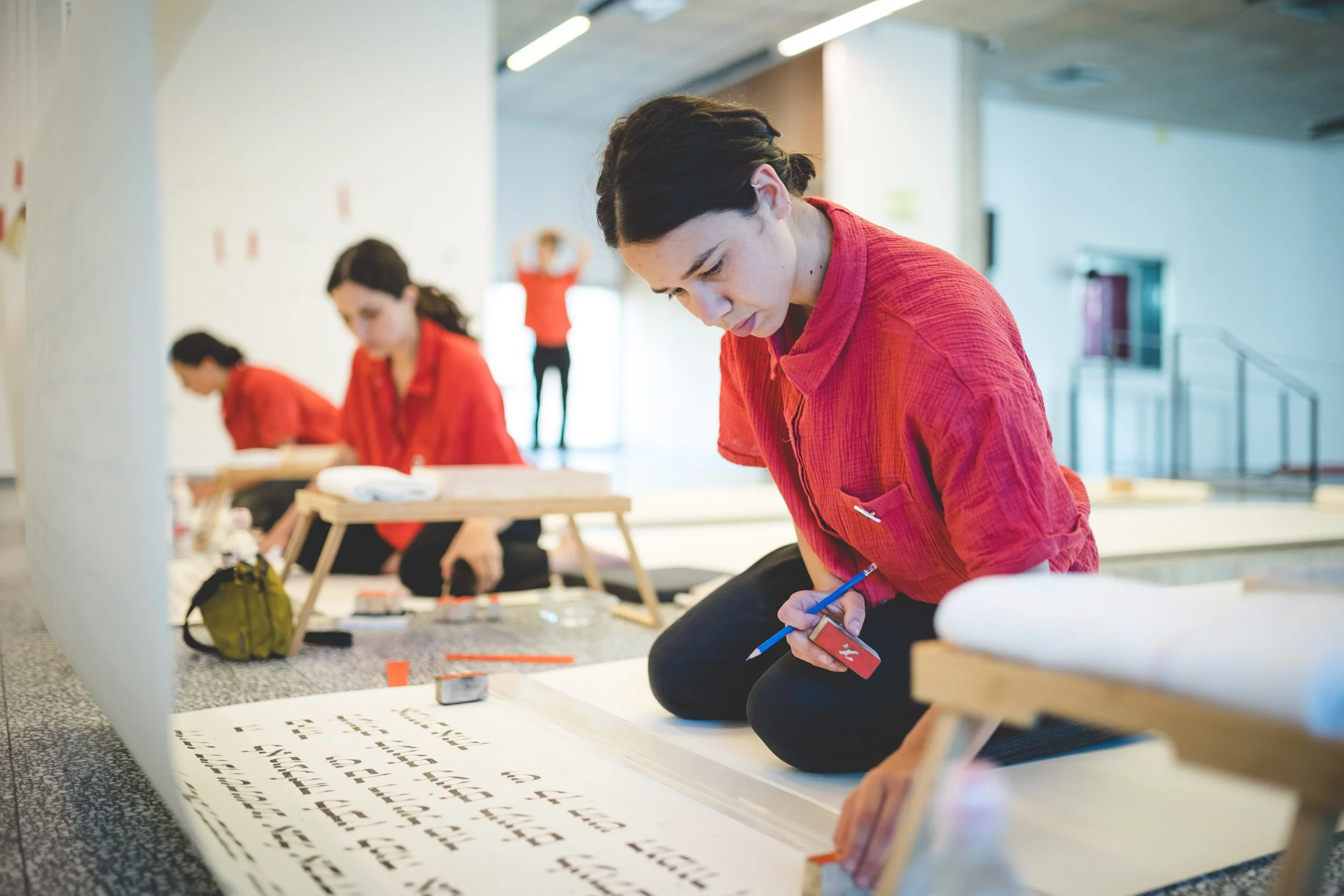 A woman in a red shirt kneels on the floor, working on a large sheet of paper with printed black text or characters. In the background, two other women in red shirts are also working on similar projects, with one woman standing and the other sitting.