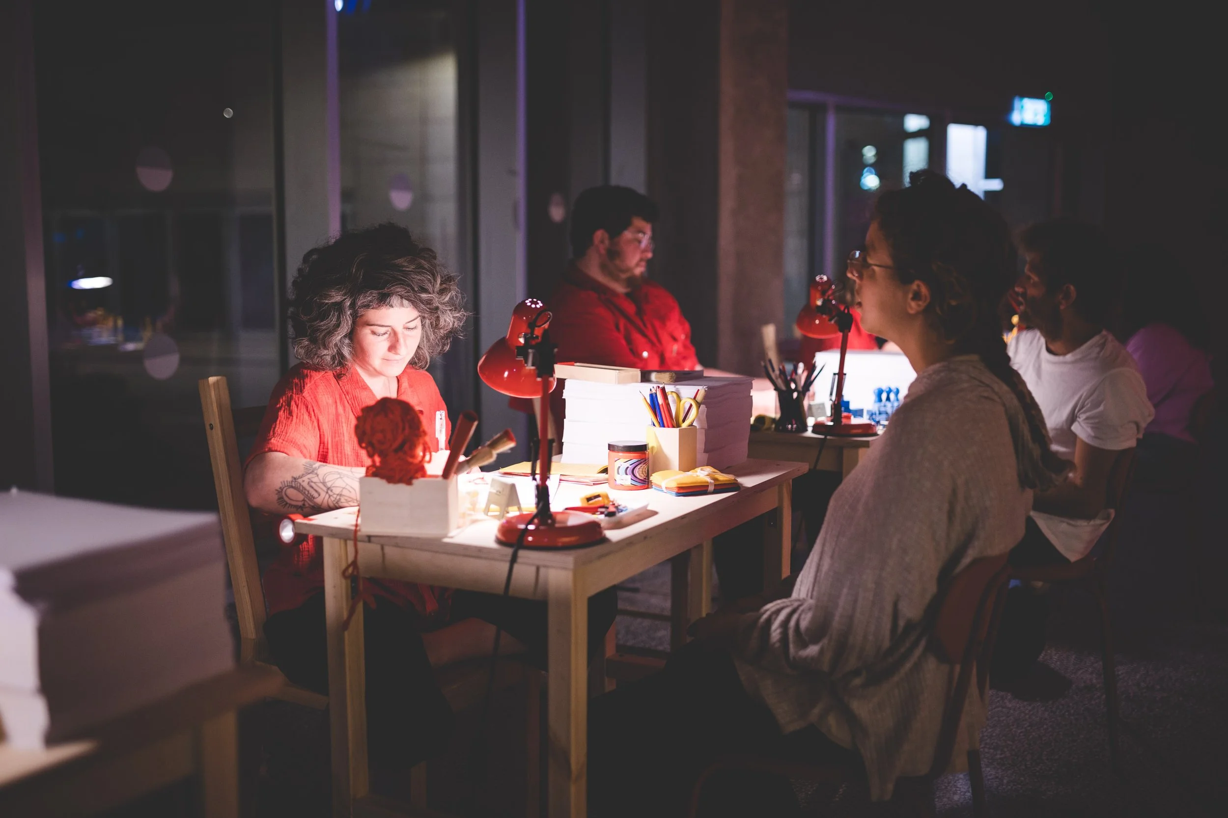 People sitting at a table in a dimly lit workspace or office, engaged in a meeting or discussion, with office supplies and lamps on the table.